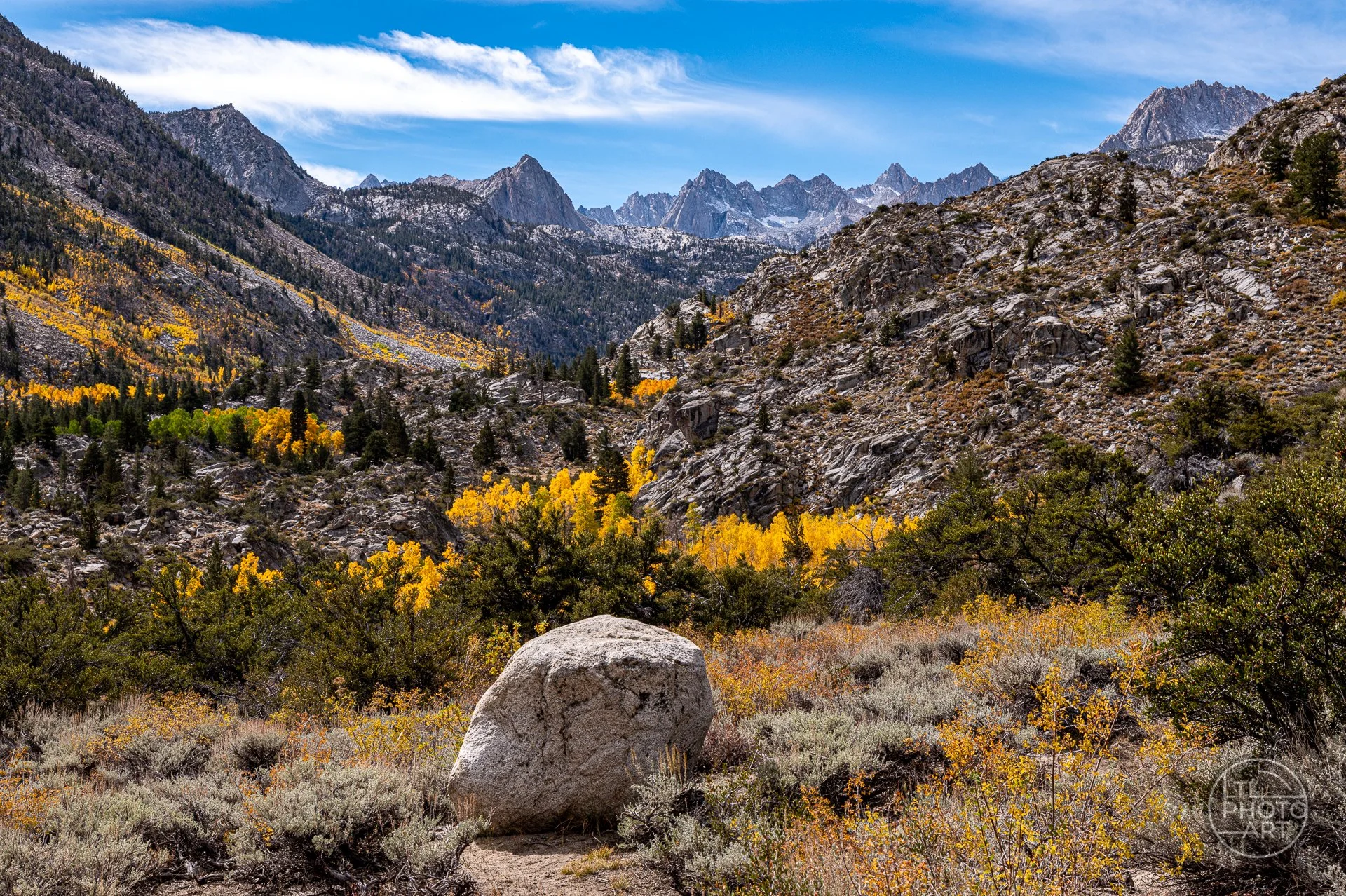 Autumn landscape along the road to North Lake in Bishop Creek Canyon, featuring golden aspen trees, rocky mountain slopes, a large foreground boulder, and distant Sierra Nevada peaks beneath a blue sky.