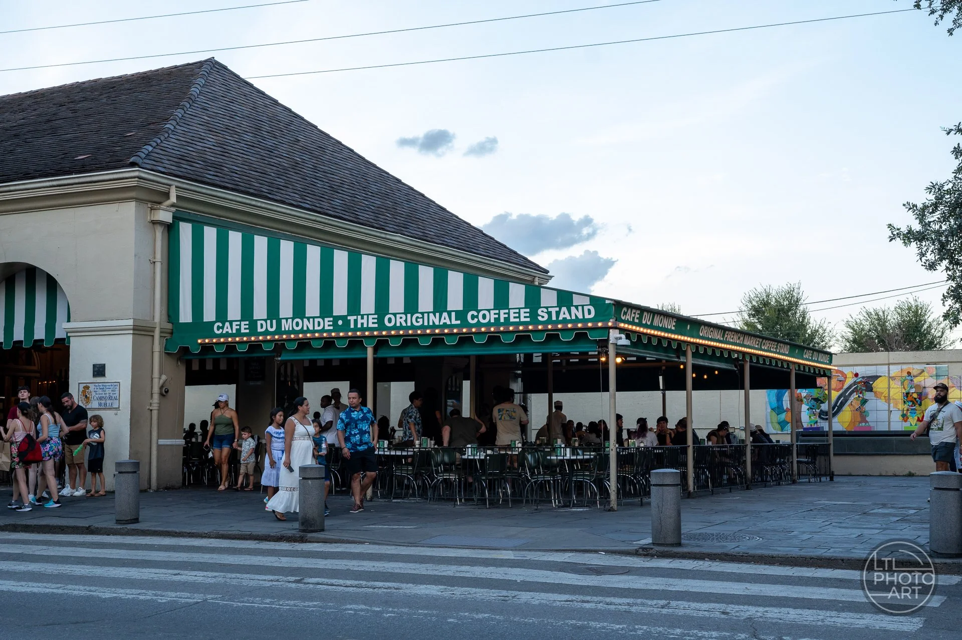 Café du Monde in New Orleans with green-and-white awnings, outdoor seating, visitors, and evening light near the French Market.