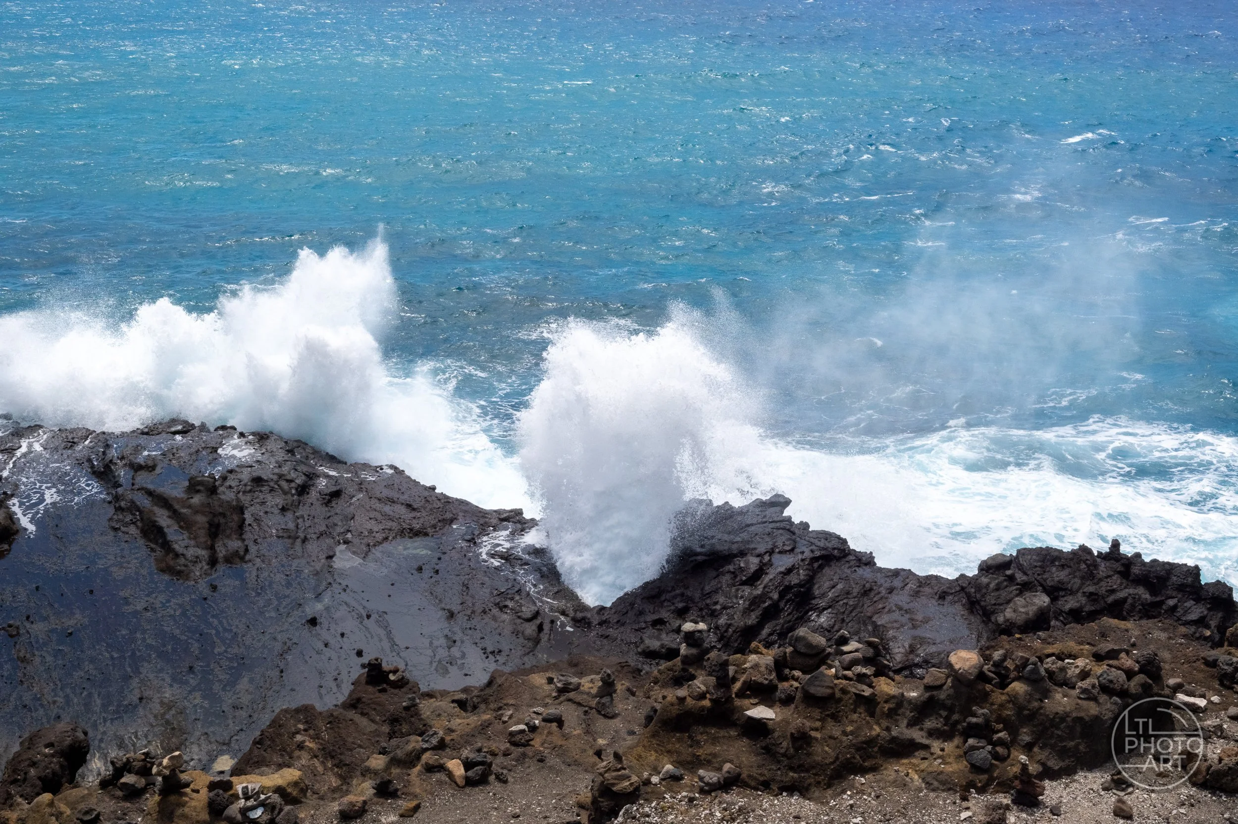 Ocean waves crash against the volcanic shoreline at Halona Blowhole on Oahu, sending white spray above the dark rocks.