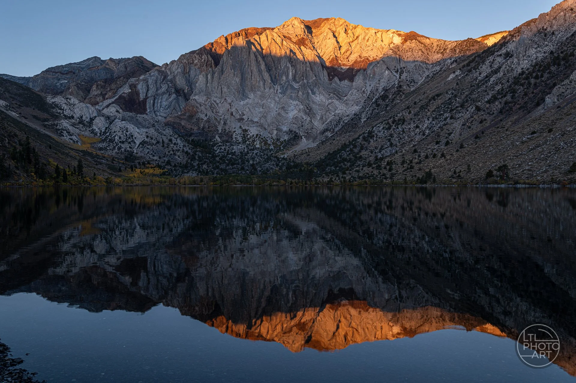 Sunrise at Convict Lake in California's Eastern Sierra, with golden sunlight illuminating the mountain peaks above the lake and reflecting in the still water beneath shadowed granite cliffs.
