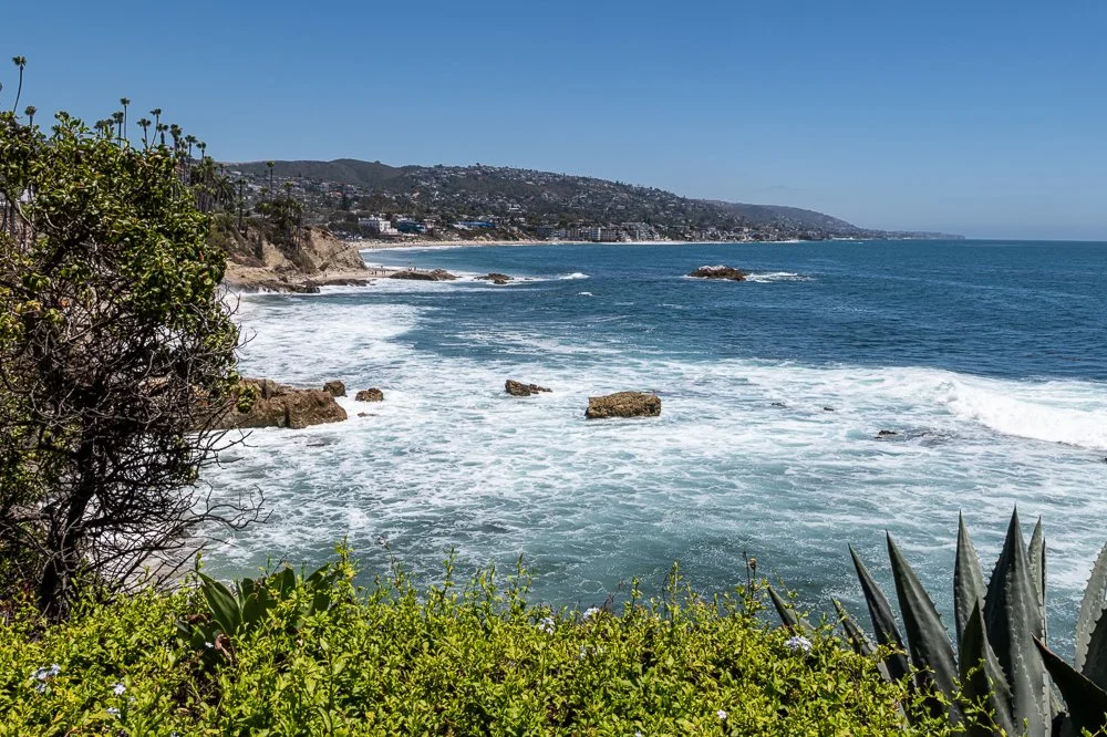 View from Heisler Park looking north toward Main Beach in Laguna Beach, California, featuring the Pacific coastline, rocky shoreline, and rolling ocean waves on a sunny day.