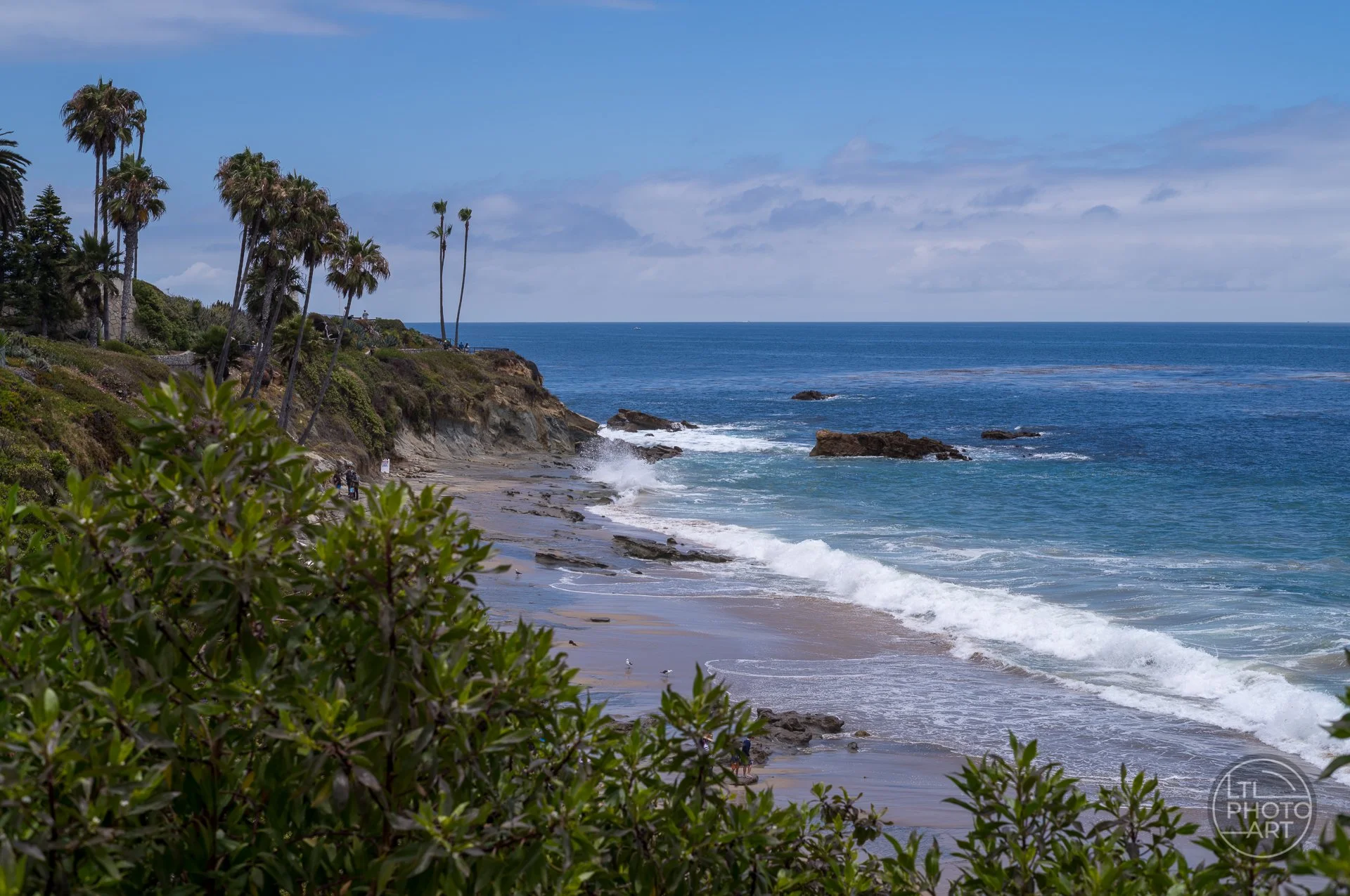 View from Heisler Park overlooking Rockpile Beach toward Monument Point in Laguna Beach, with waves, cliffs, palm trees, and the Pacific Ocean.