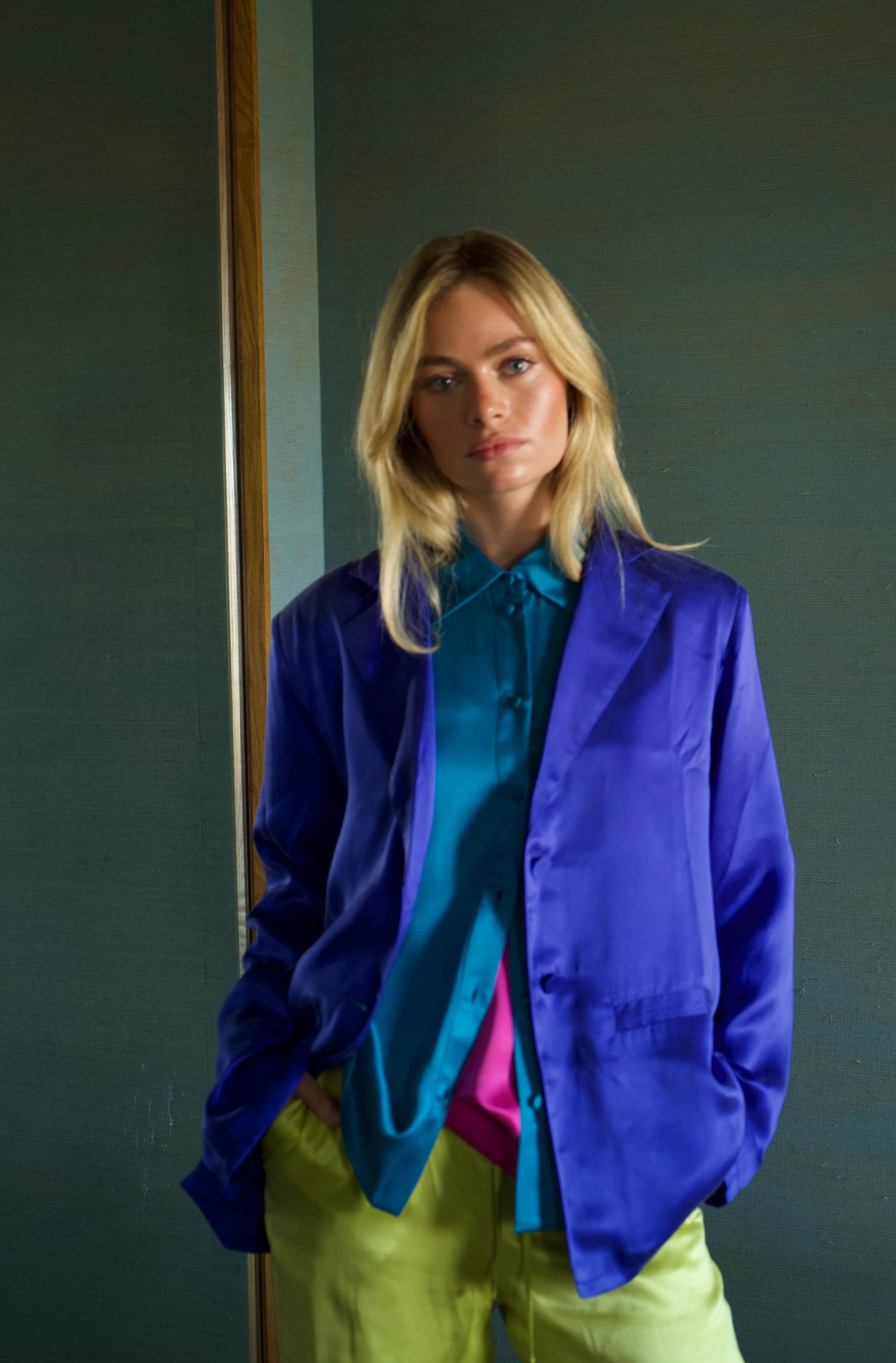 A young woman with blonde hair wearing a shiny purple jacket, a teal shirt underneath, and yellow pants stands against a plain green wall.