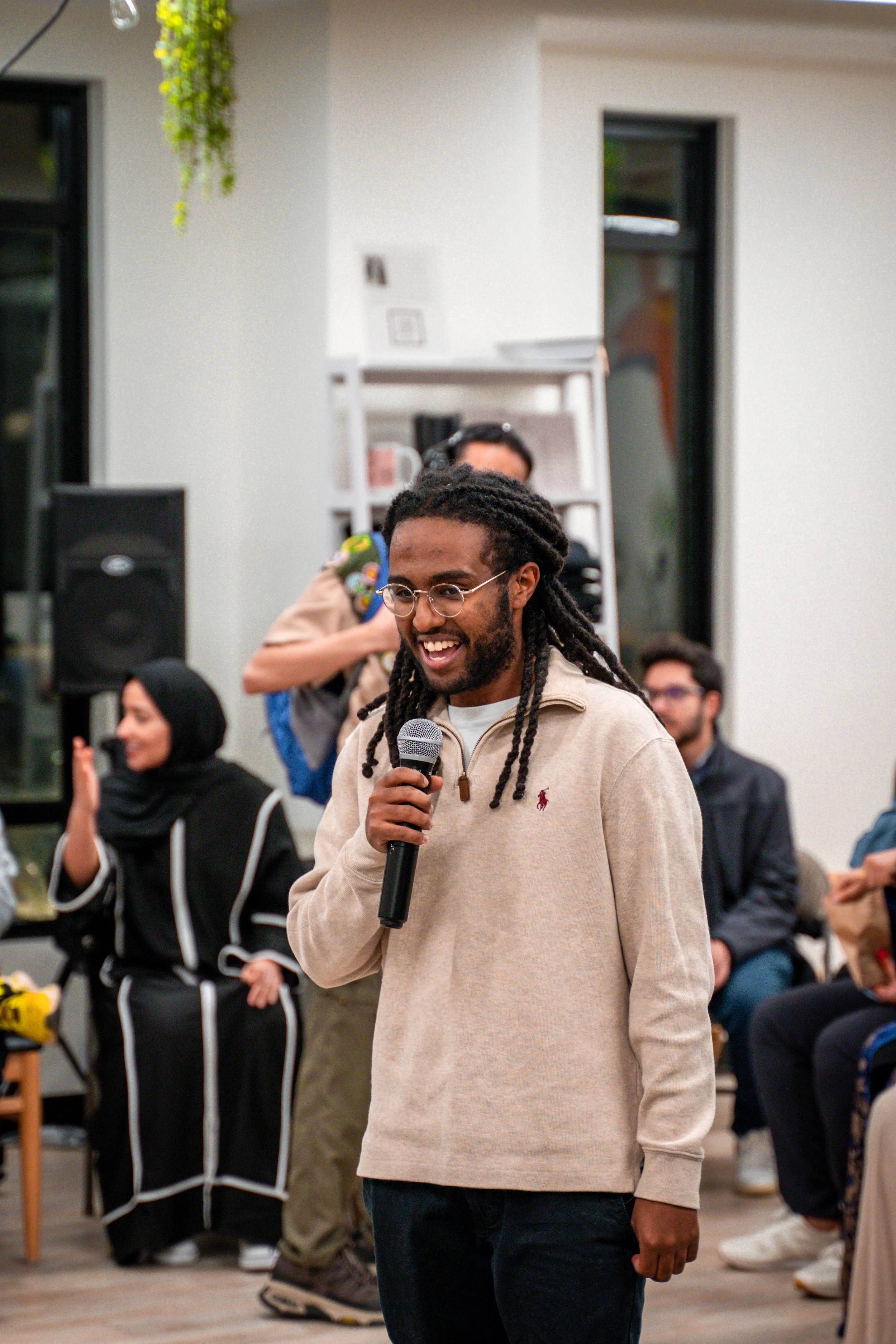 A man with glasses and dreadlocks is smiling and holding a microphone in a room with seated people in the background. He is wearing a beige sweater with a logo.
