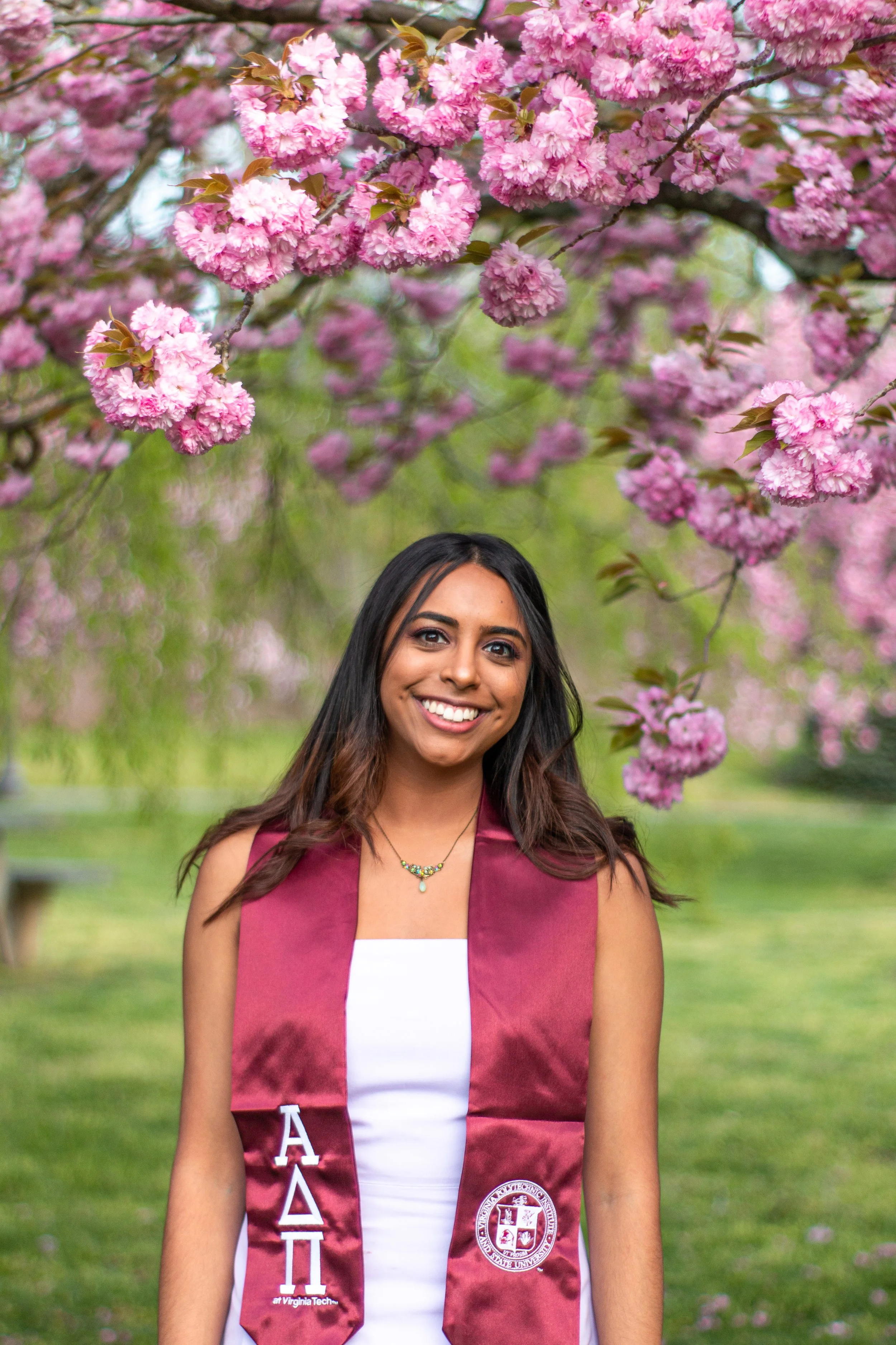 Young woman in graduation attire standing in front of blossoming pink cherry blossoms.
