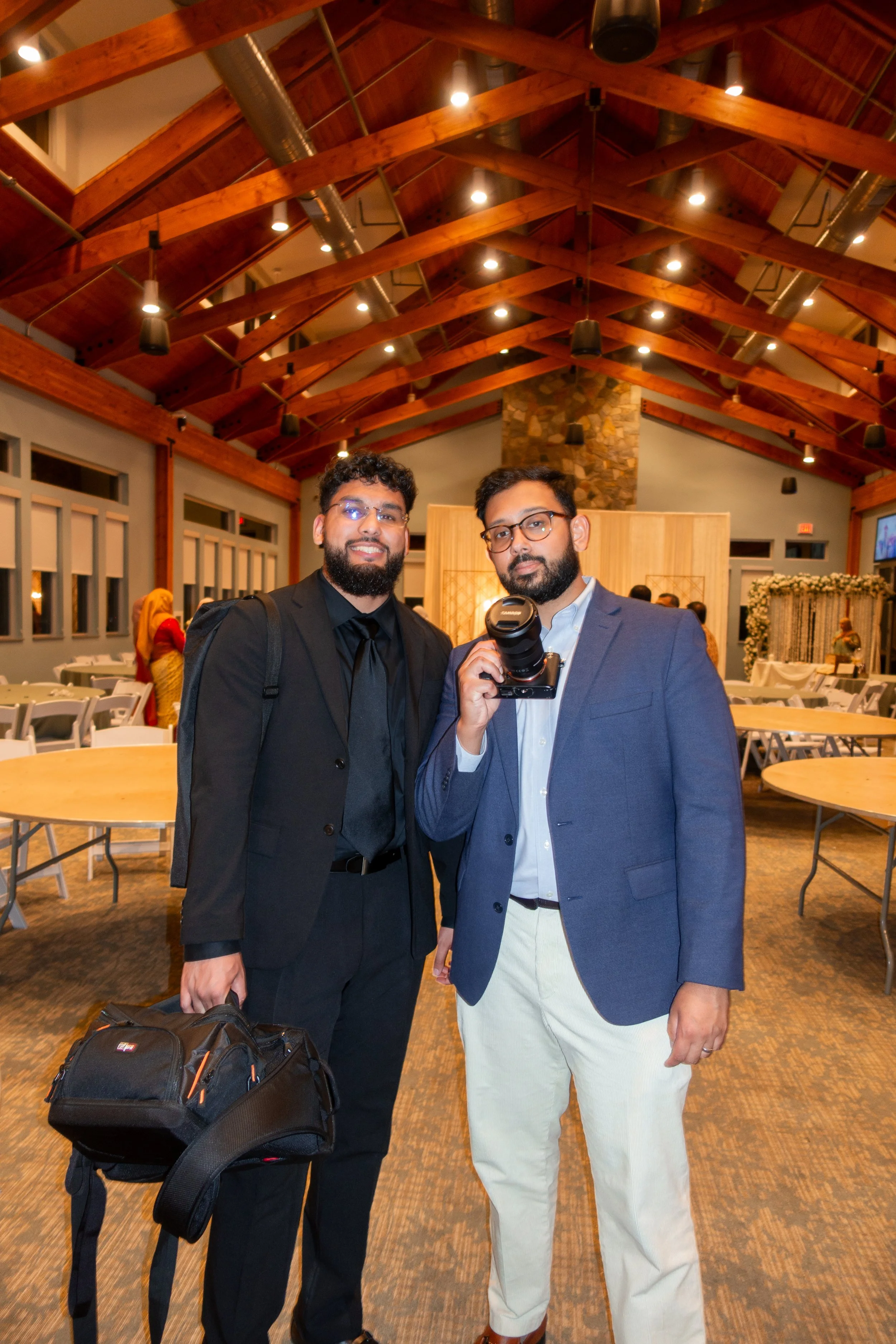 Two men dressed in formal attire standing inside a decorated event hall with a high wooden ceiling and tables, one holding a camera, both smiling at the camera.