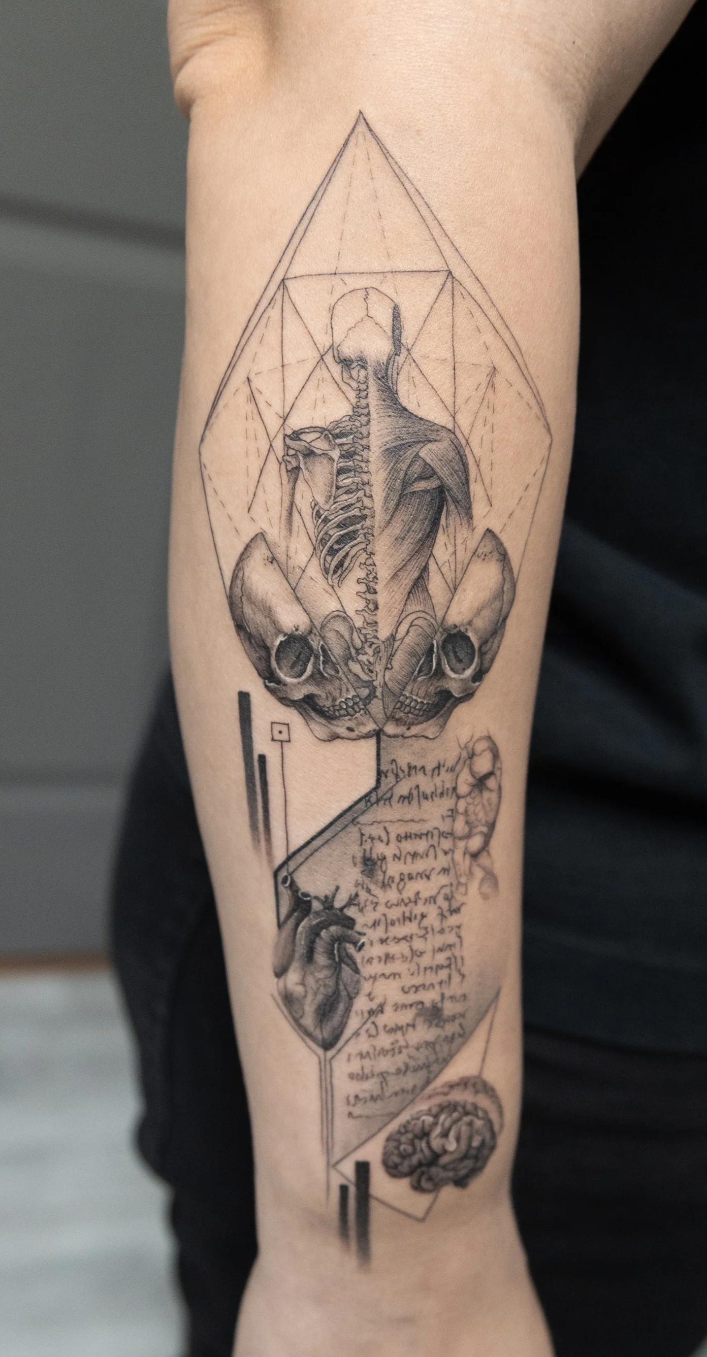 Tattoo of anatomical sketches including a human skeleton and skulls, a heart, a brain, geometric shapes, and handwritten notes.