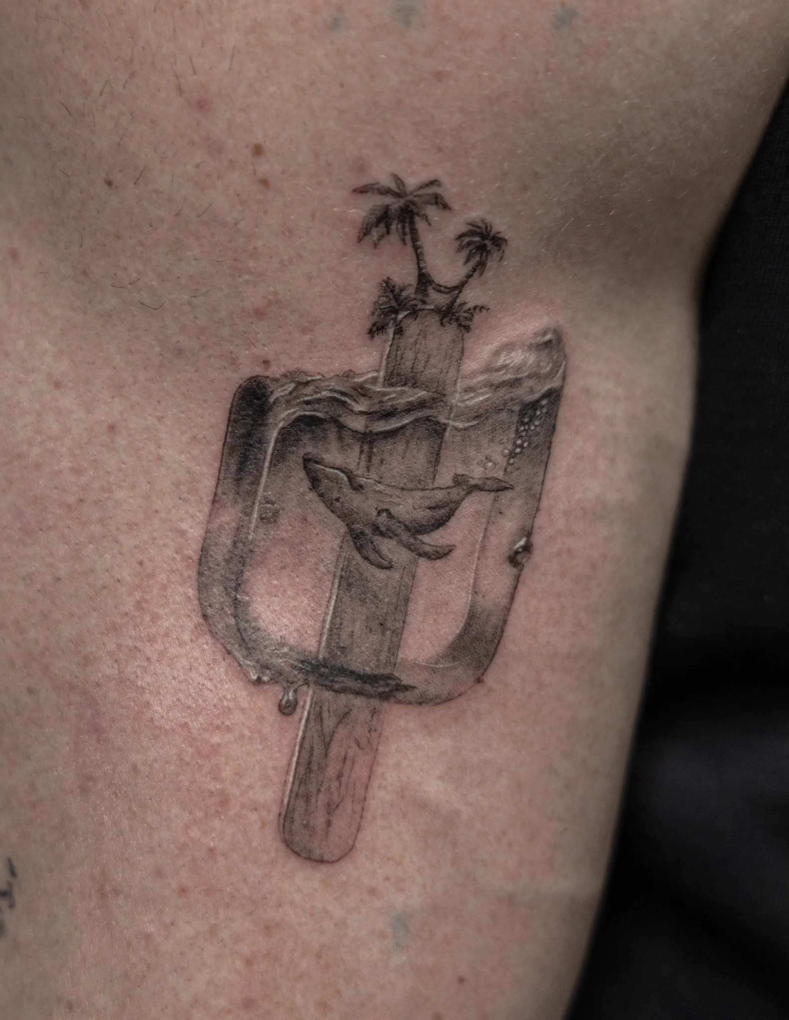 Tattoo of a popsicle with a whale inside and a small beach scene with palm trees on top of the popsicle stick.