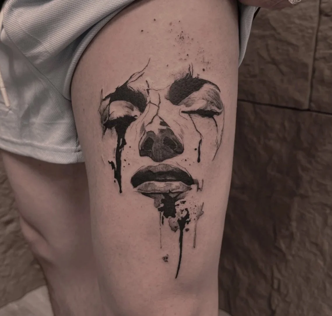 A black and gray tattoo of a woman's face with closed eyes and tears, featuring a watercolor style with drips and splashes, on a person's thigh.