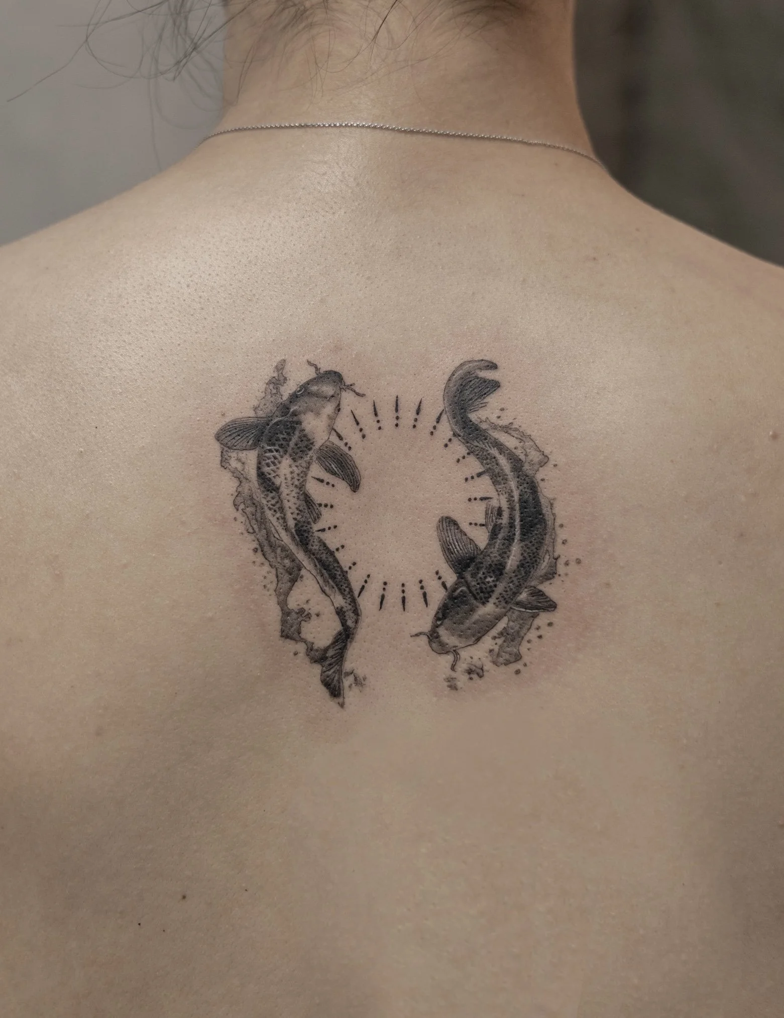 Tattoo of two koi fish swimming in a circle with small dots and lines around them on a person's upper back.