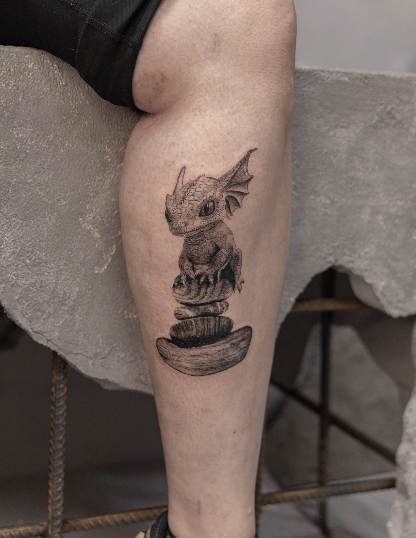 Tattoo of a baby dragon with big eyes and small wings, sitting on a rock stack, on a person's leg.