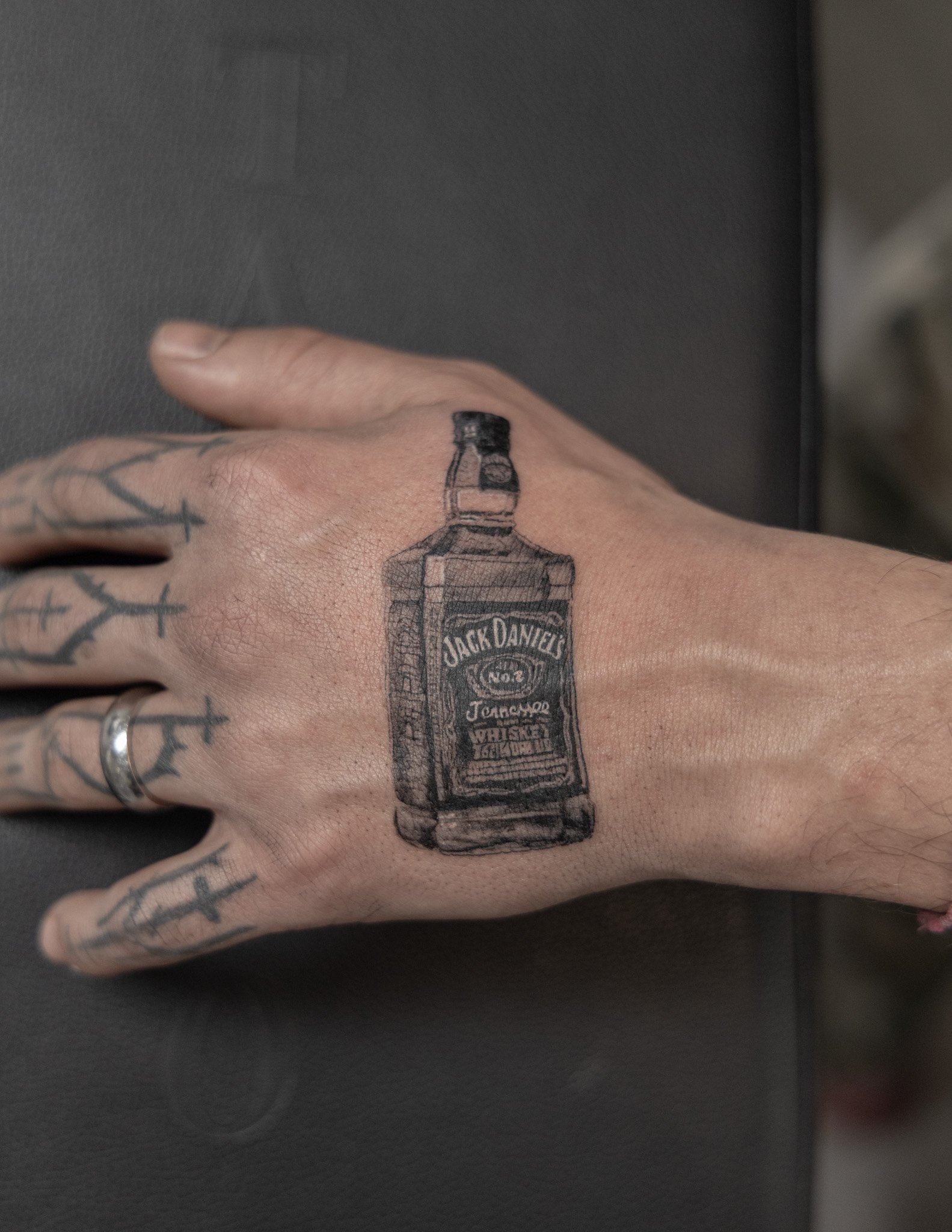 Tattoo of Jack Daniel's whiskey bottle on a person's hand, with additional tattoos of crosses and tattoos on fingers.