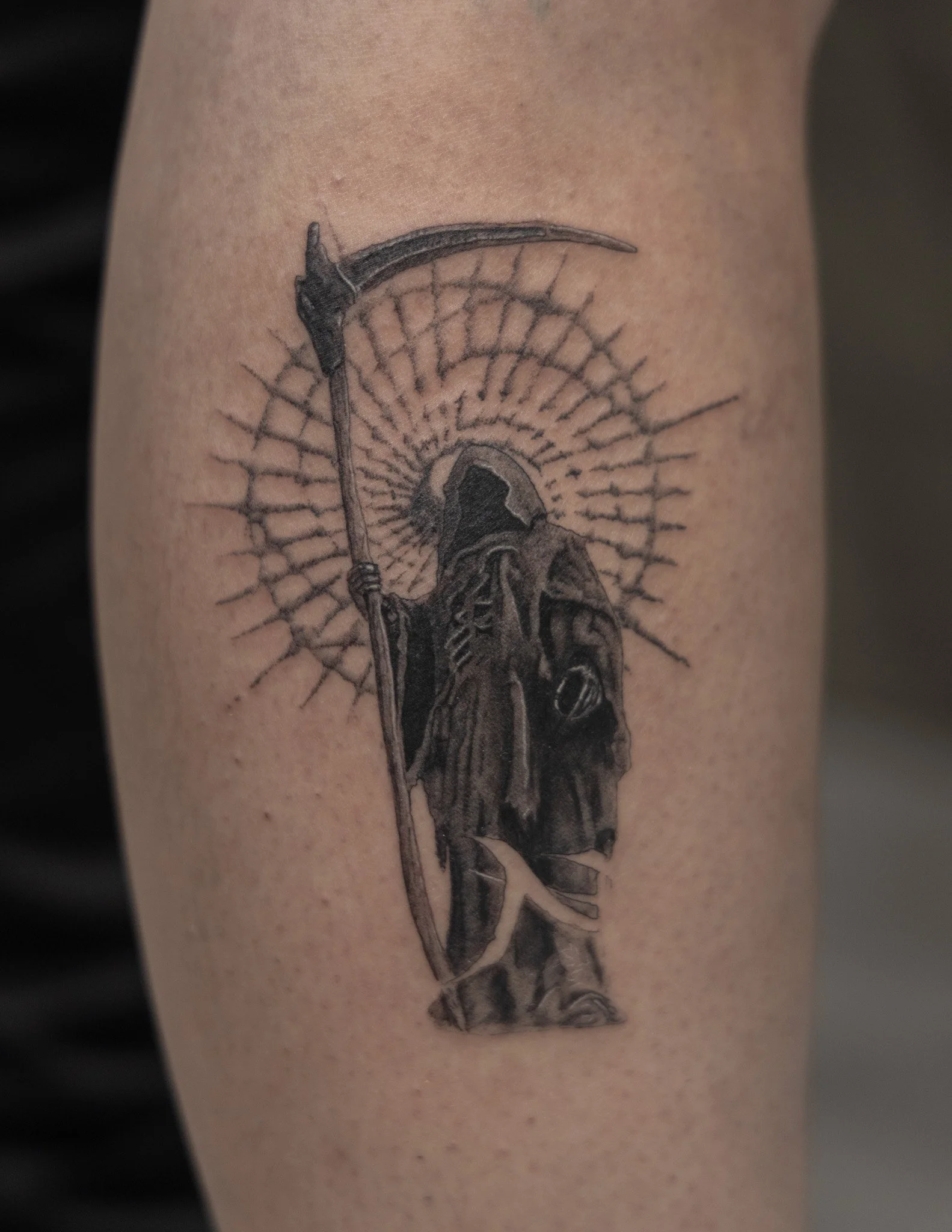Black and grey tattoo of the Grim Reaper holding a scythe, with a radiant halo behind its head.