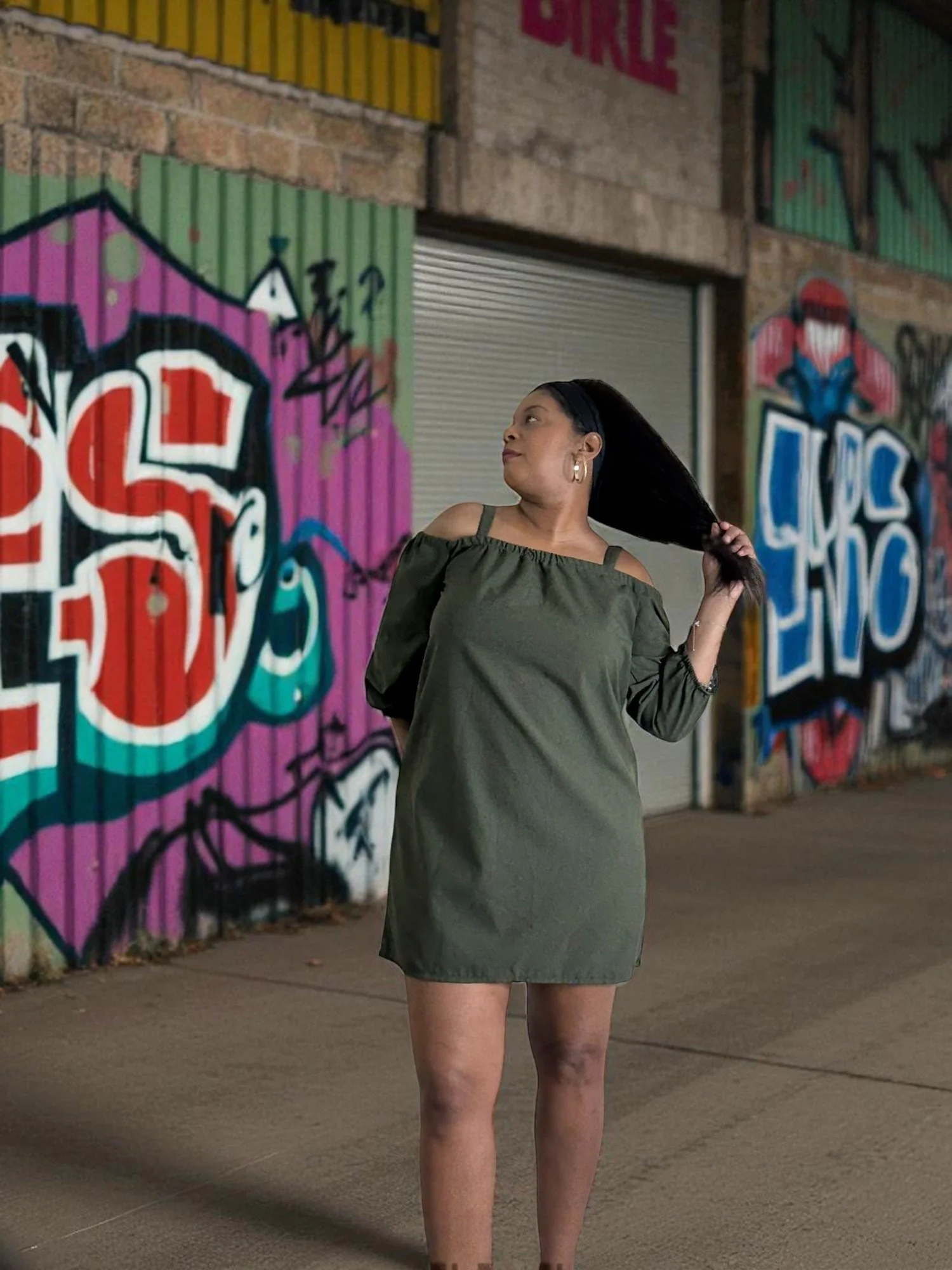 A woman in an olive green dress stands in front of a colorful graffiti wall at night, holding her long black hair with her right hand.