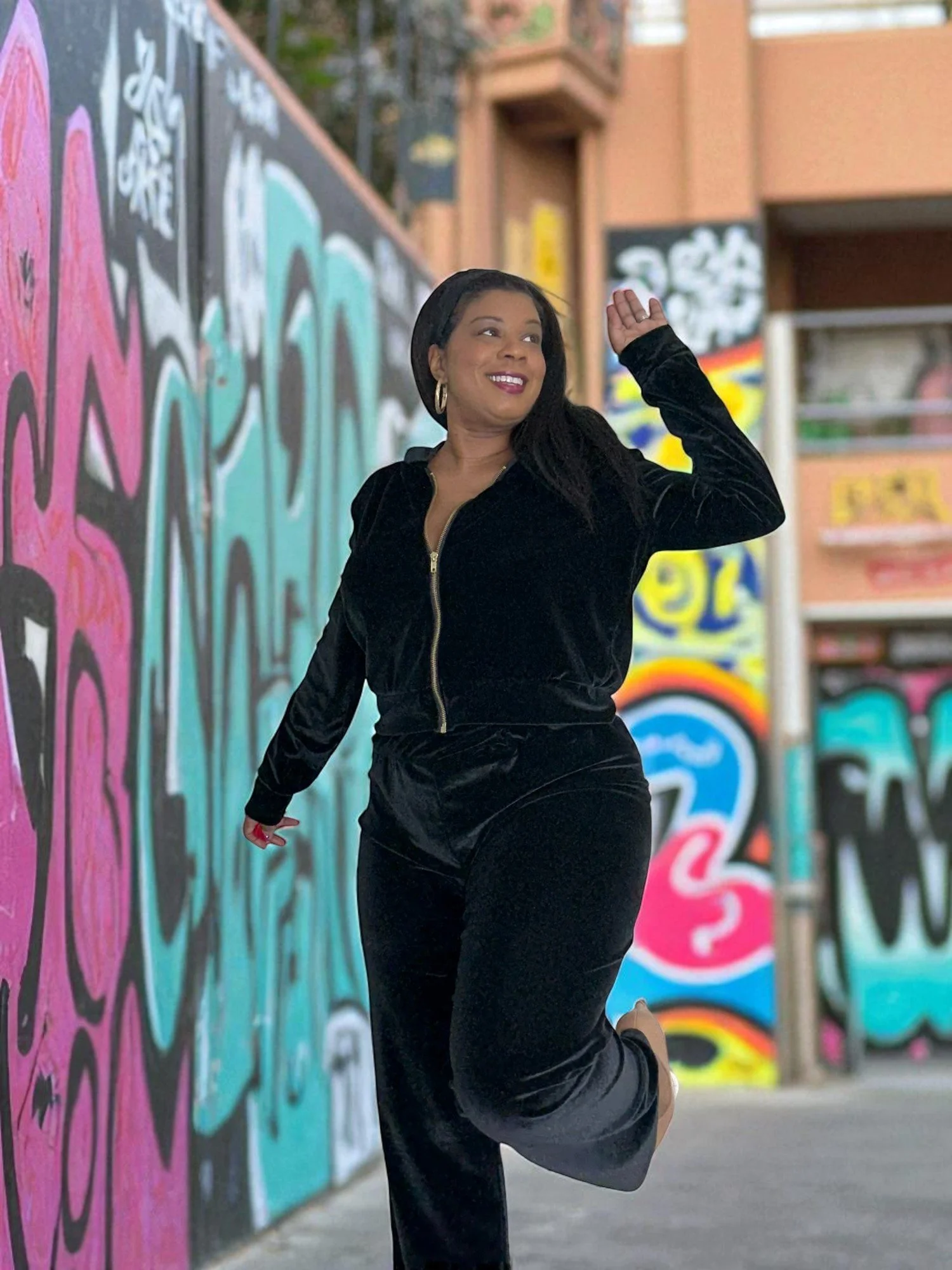A woman in a black velvety tracksuit smiling and posing in front of a colorful graffiti wall in an urban alleyway.