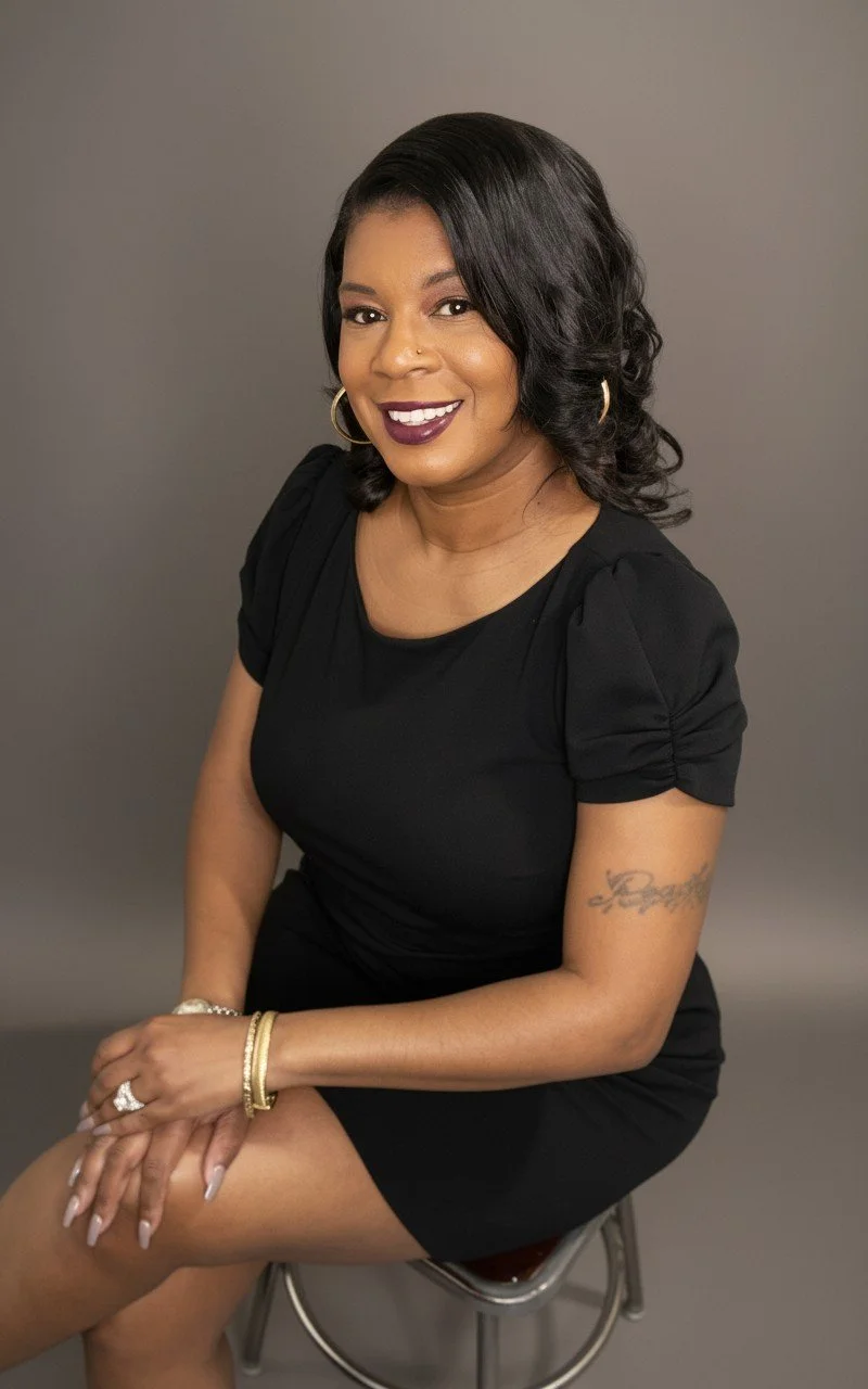 A woman sitting on a stool, smiling, wearing a black dress with puffy sleeves, gold hoop earrings, and bracelets, with a gray background.