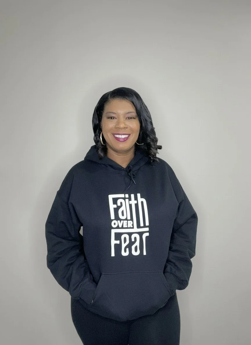 A woman smiling, wearing a black hoodie with the phrase "Faith Over Fear" printed on it, standing against a plain light-colored wall.