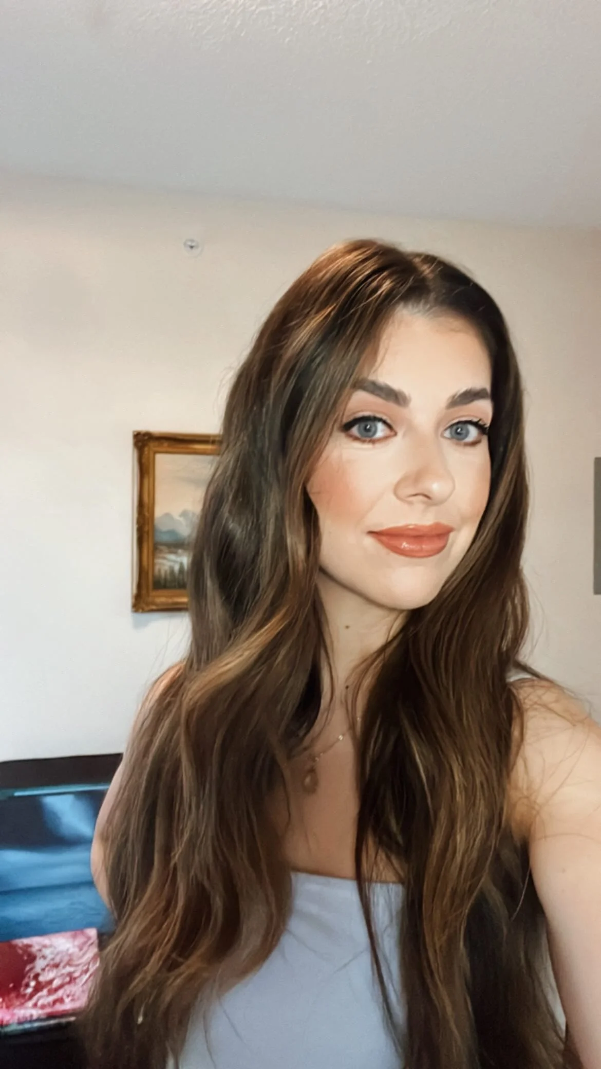 Melissa, tarot reader and founder of Empowered Gems Tarot, smiling with long wavy brown hair, seated in front of a framed painting.