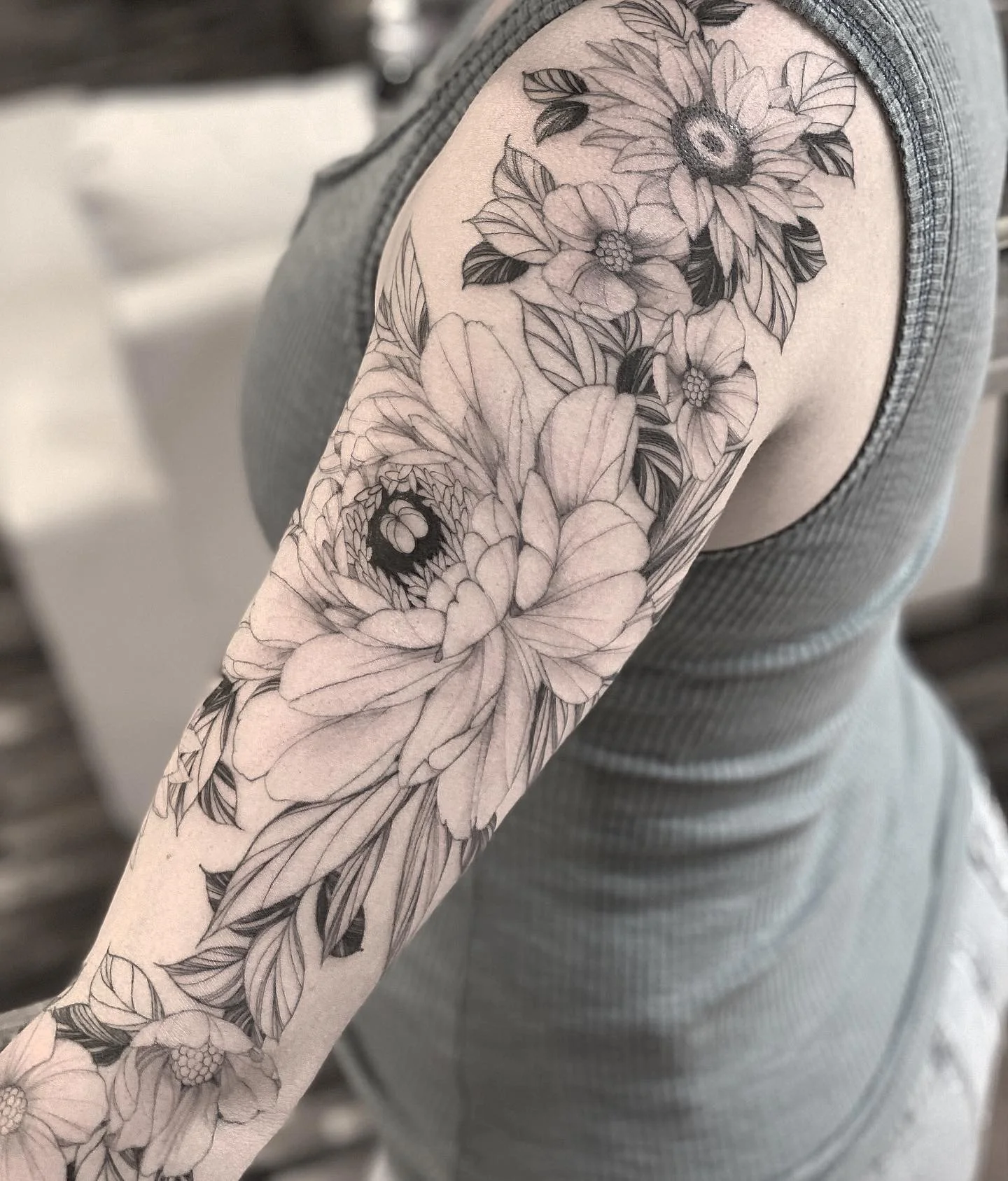 Black and gray floral tattoo on person's upper arm and shoulder, featuring sunflowers, peonies, and leaves.