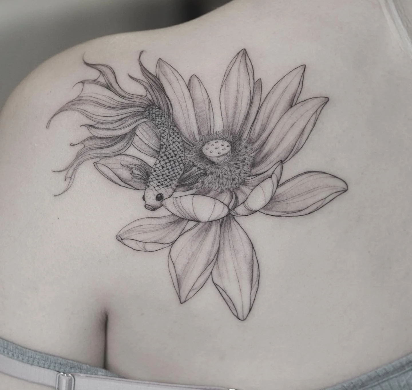 A black and white tattoo of a fish, resembling a betta, swimming among large sunflower petals on a person's chest.