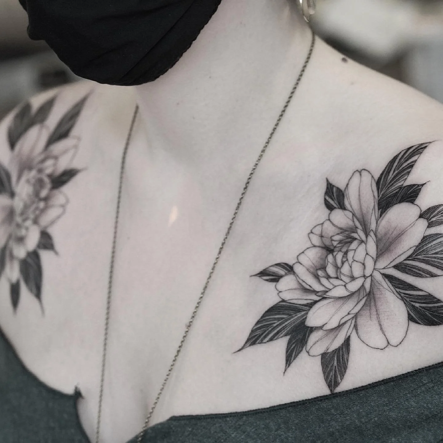 Close-up of a person's shoulder and chest with detailed floral tattoos, wearing a watch and a necklace, with a black face mask covering part of their face.