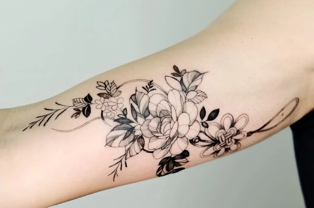 Black outline floral tattoo on inner forearm with a large blooming flower, leaves, and a small decorative element resembling a flower on the right.