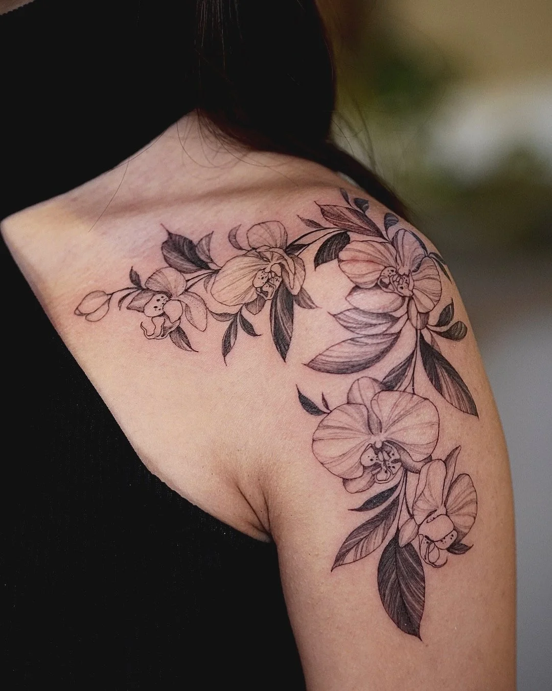 Black and gray floral tattoo on person's shoulder and upper arm, including orchids and leaves.