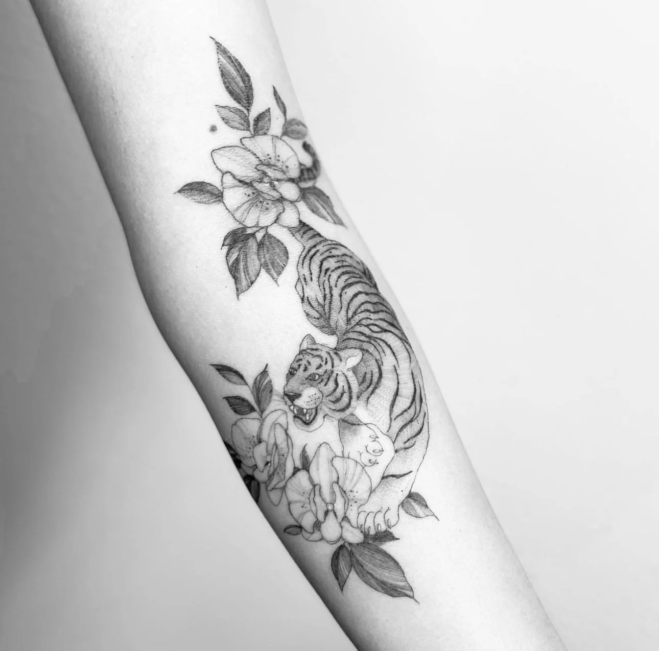 Black and white tattoo of a tiger surrounded by flowers and leaves on a person's arm.