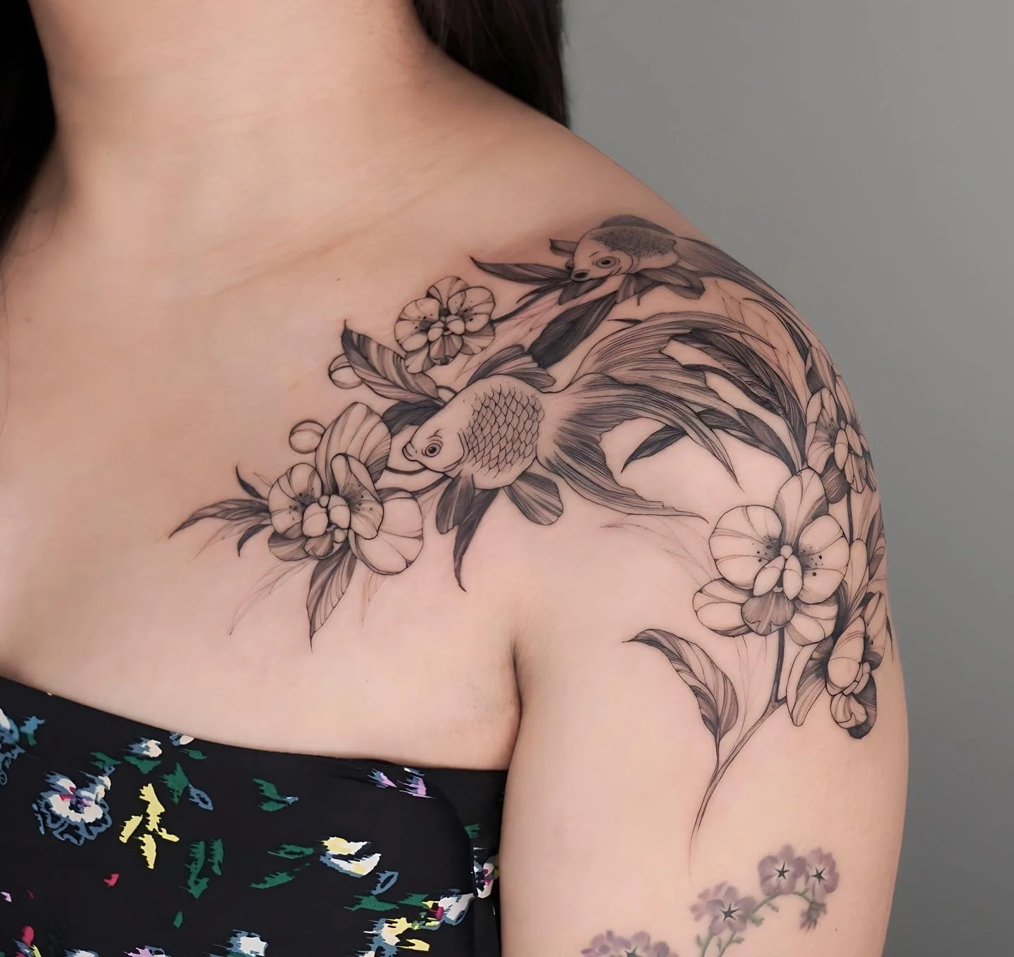A black and gray floral tattoo featuring two fish and flowers on a person's shoulder and upper arm.