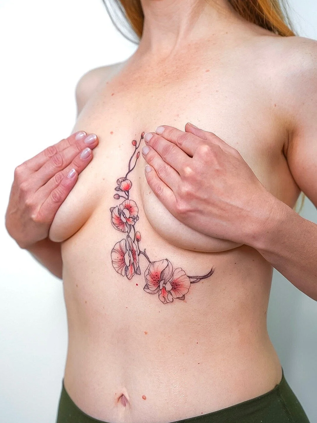 A woman with a floral tattoo on her chest, covering her breasts with her hands, wearing green pants.