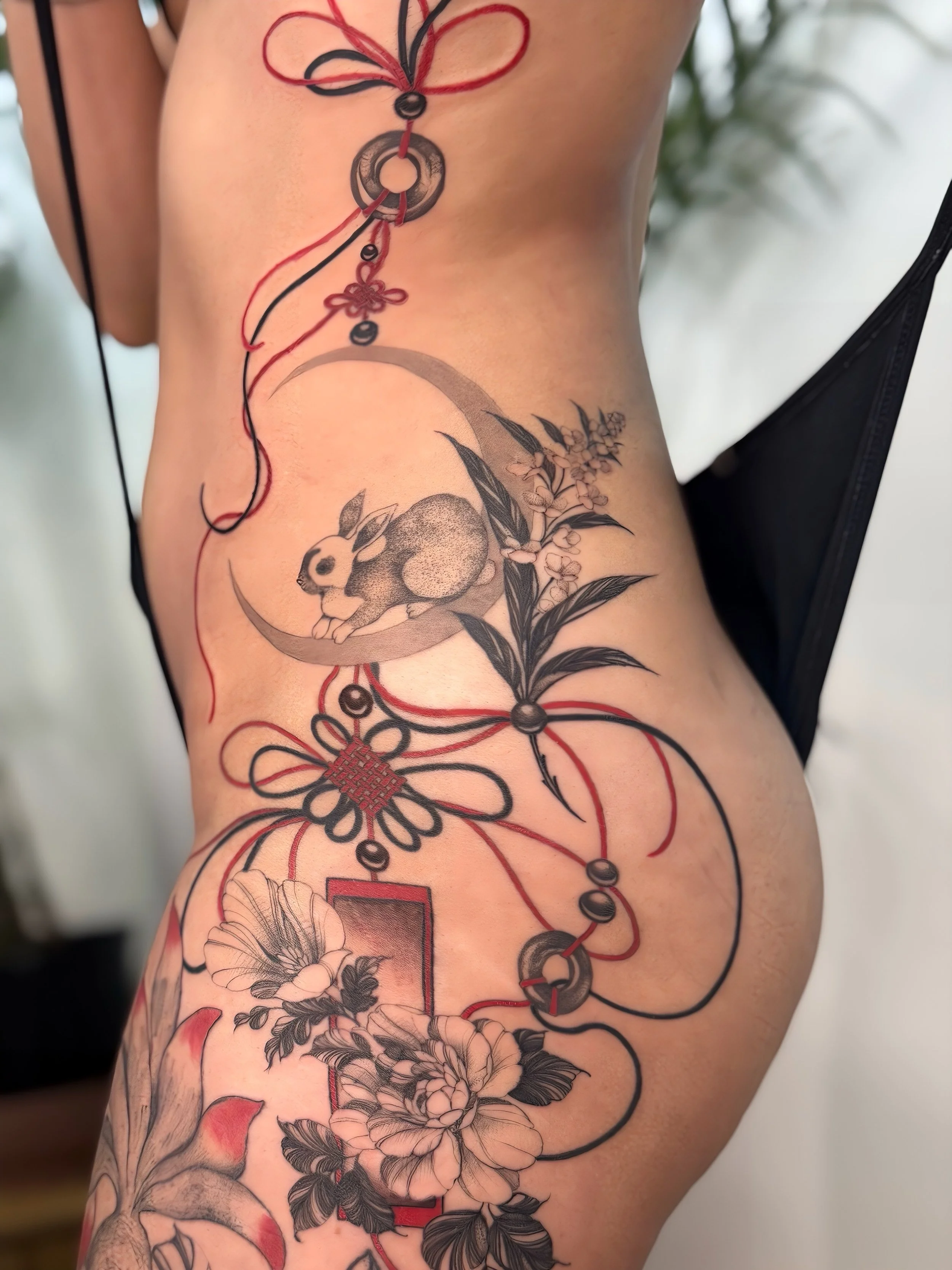 A close-up of a person's side and torso featuring a detailed black and gray tattoo of a rabbit, flowers, and geometric elements with red accents.
