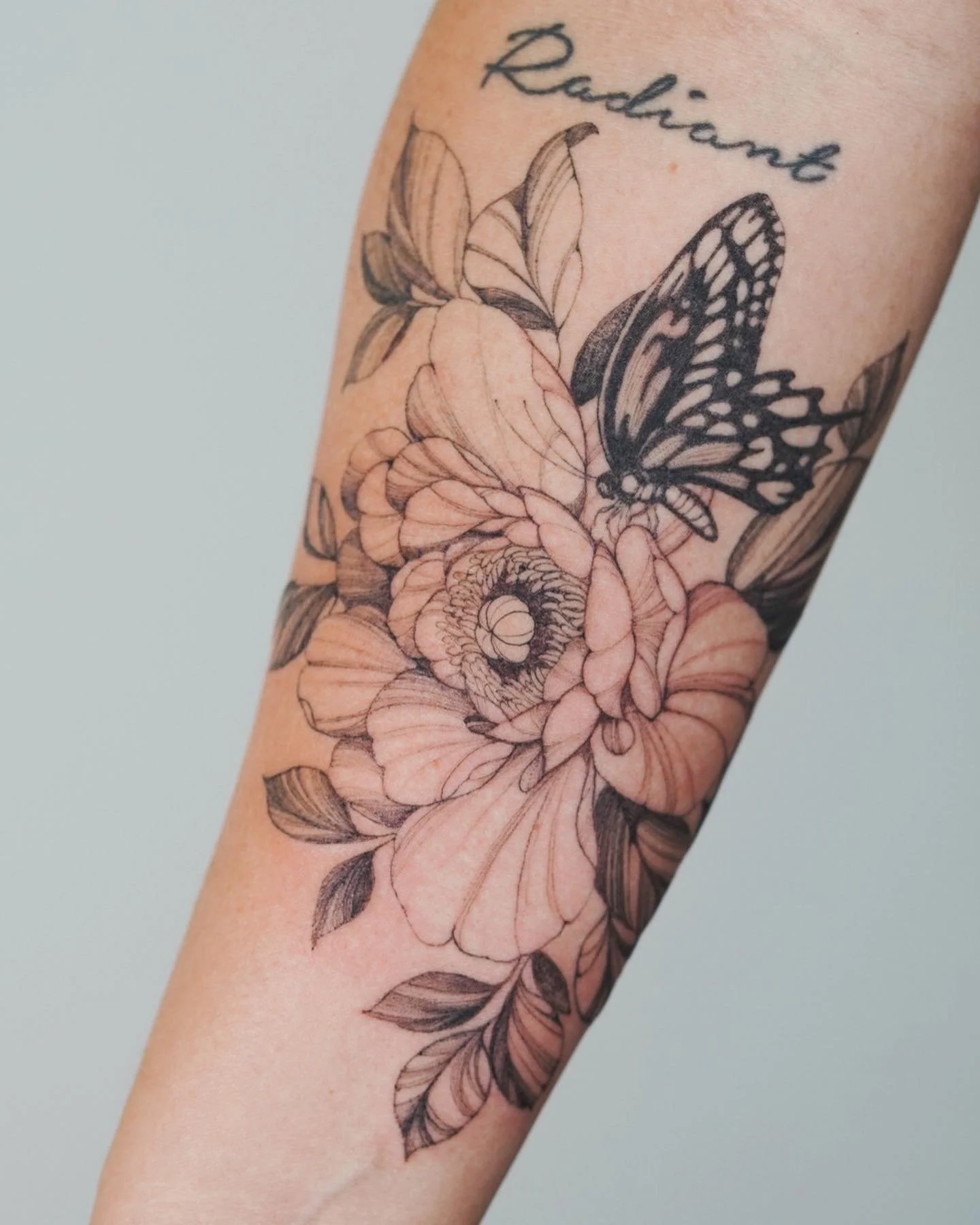 Black and gray floral tattoo with two butterflies on a person's arm, and the word 'Radiant' written above.