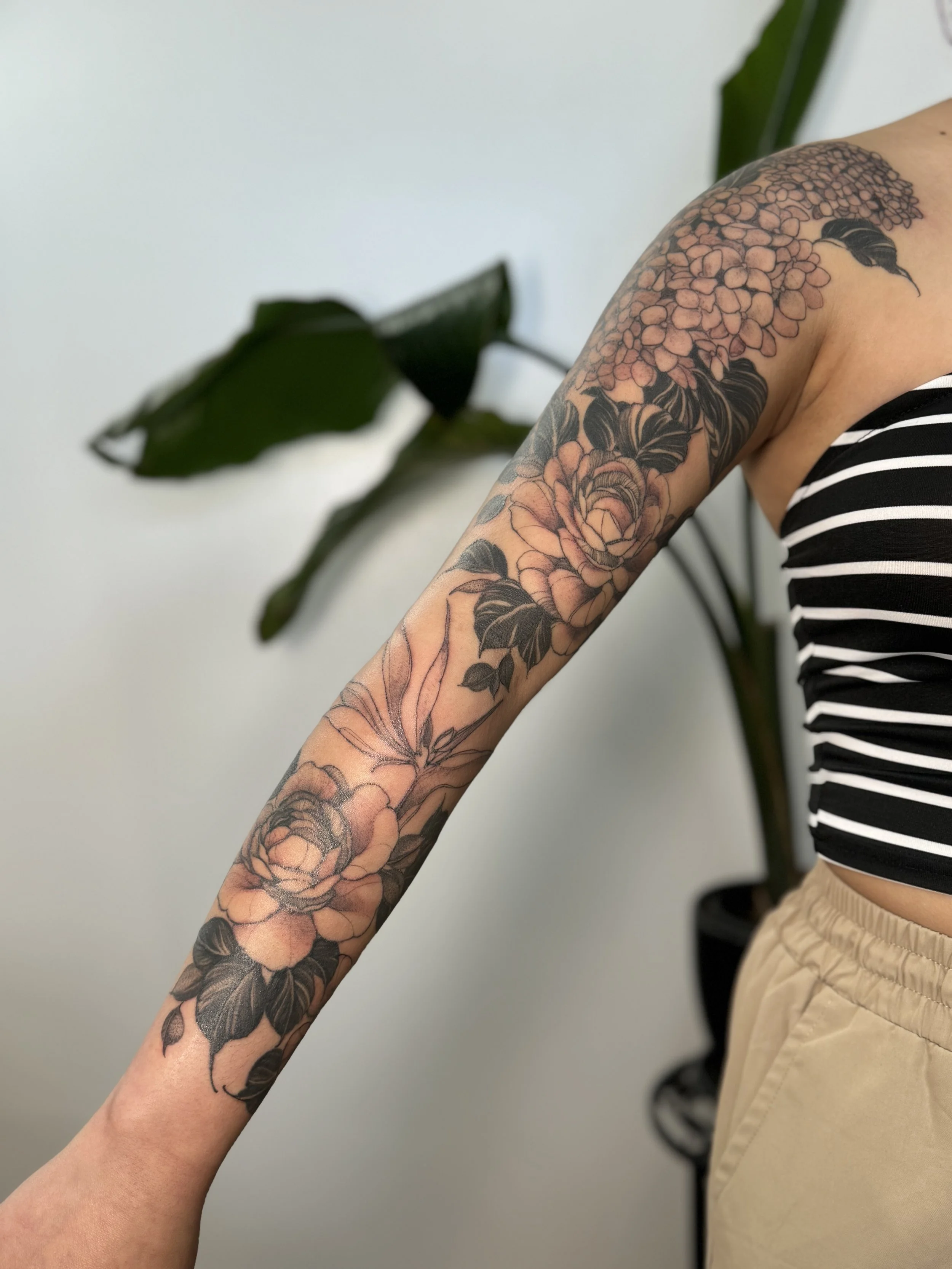 Tatted arm with floral design extending from shoulder to wrist, featuring roses, hydrangeas, and leaves in black and subtle pink colors.