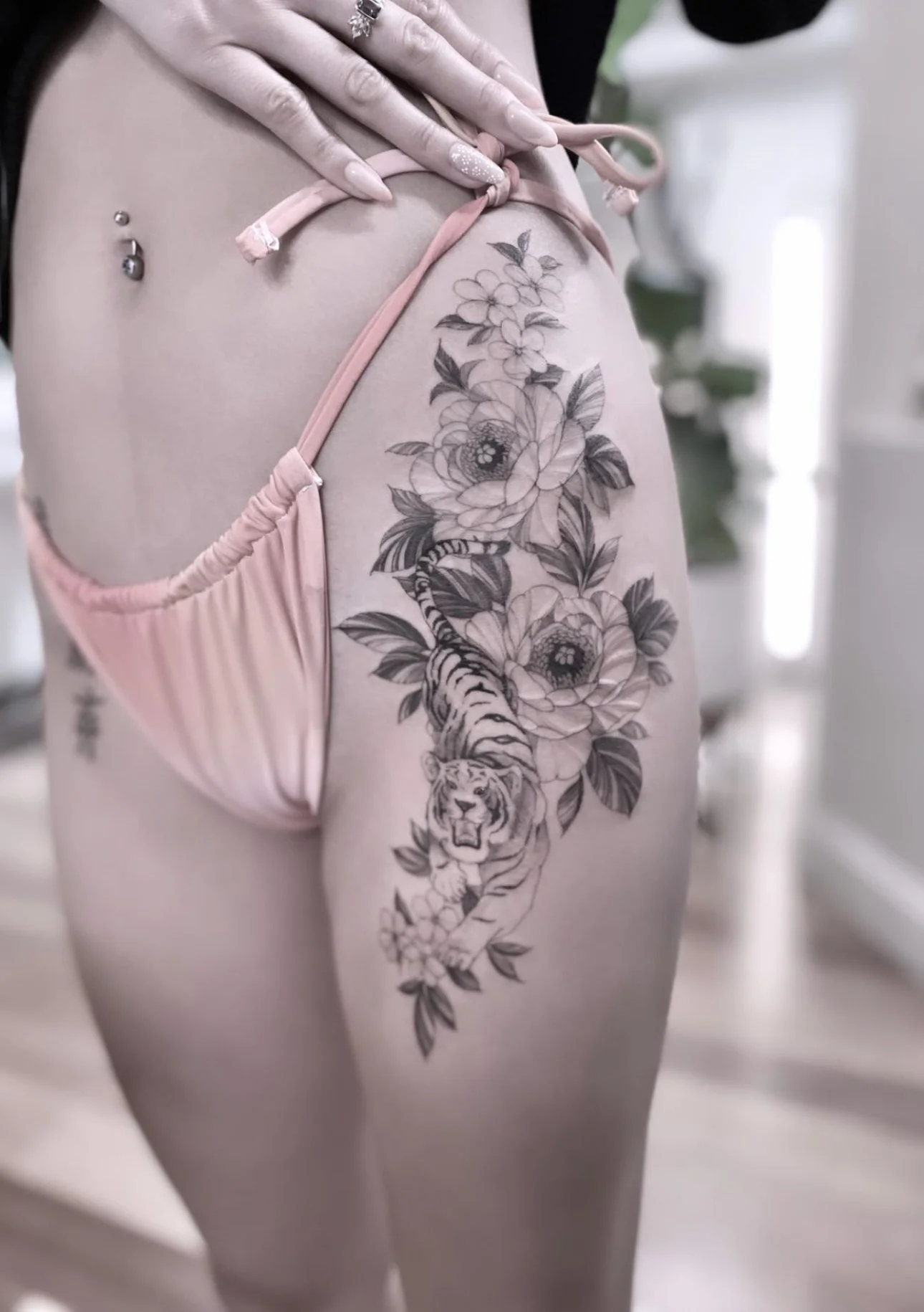 Tattoo of a lion cub surrounded by large flowers and a tiger inside a woman's upper thigh and side, with a belly button piercing and wearing pink lingerie.