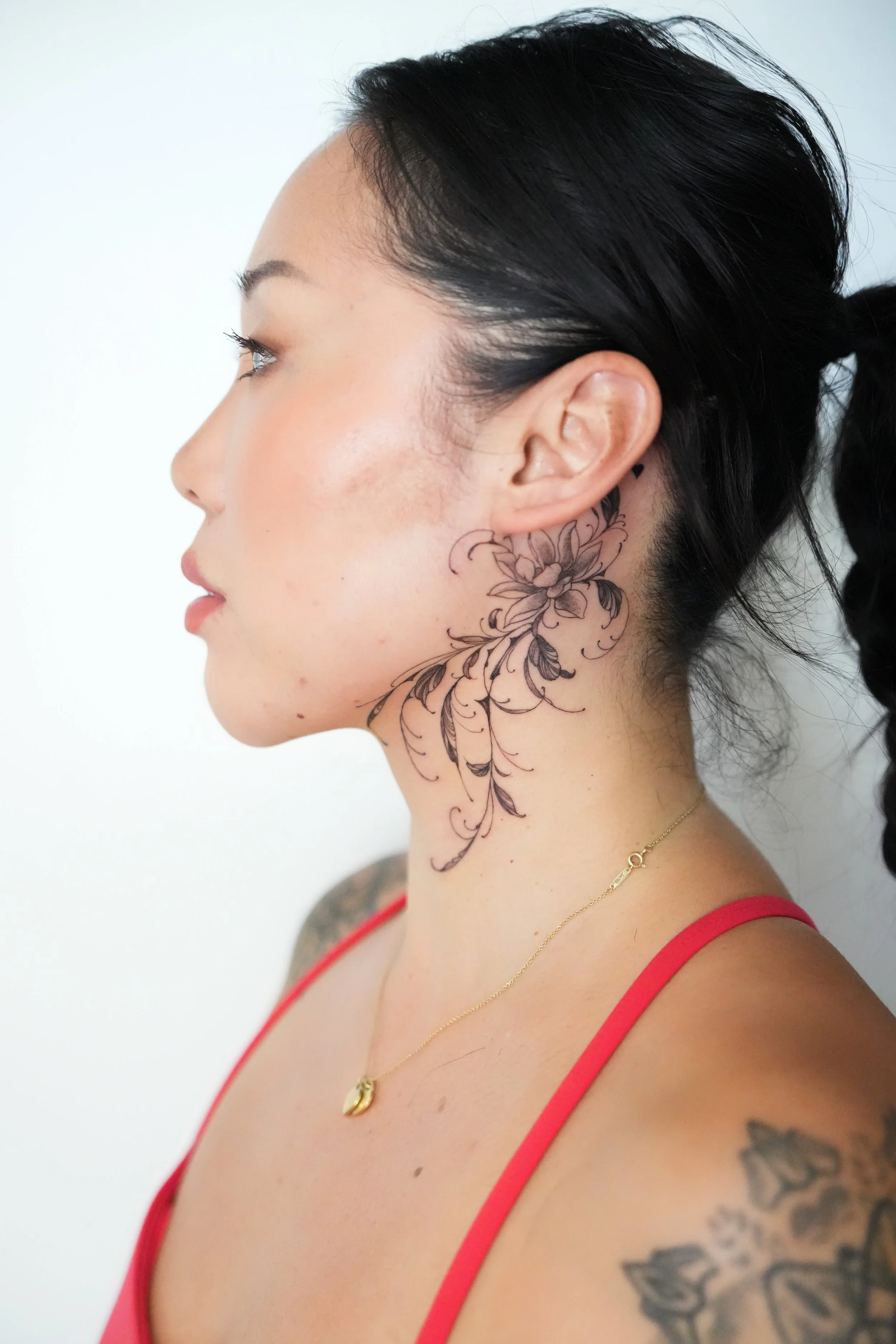 Profile of a woman with black hair, a floral tattoo on her neck, and tattoos on her shoulder, wearing a red top and a gold necklace.