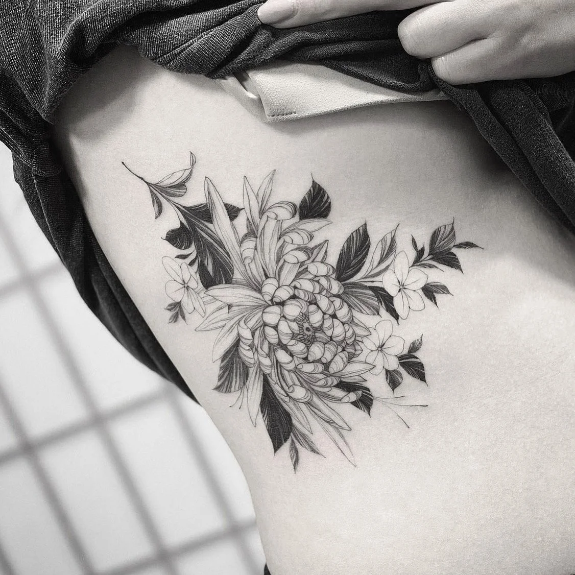 Black and gray floral tattoo on person's upper arm, featuring intricate petals and leaves in a detailed, illustrative style.