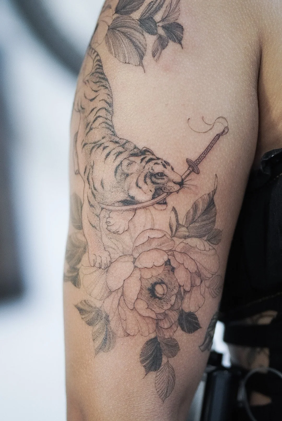 Tattoo of a tiger with a syringe in its mouth, surrounded by flowers and leaves on upper arm.