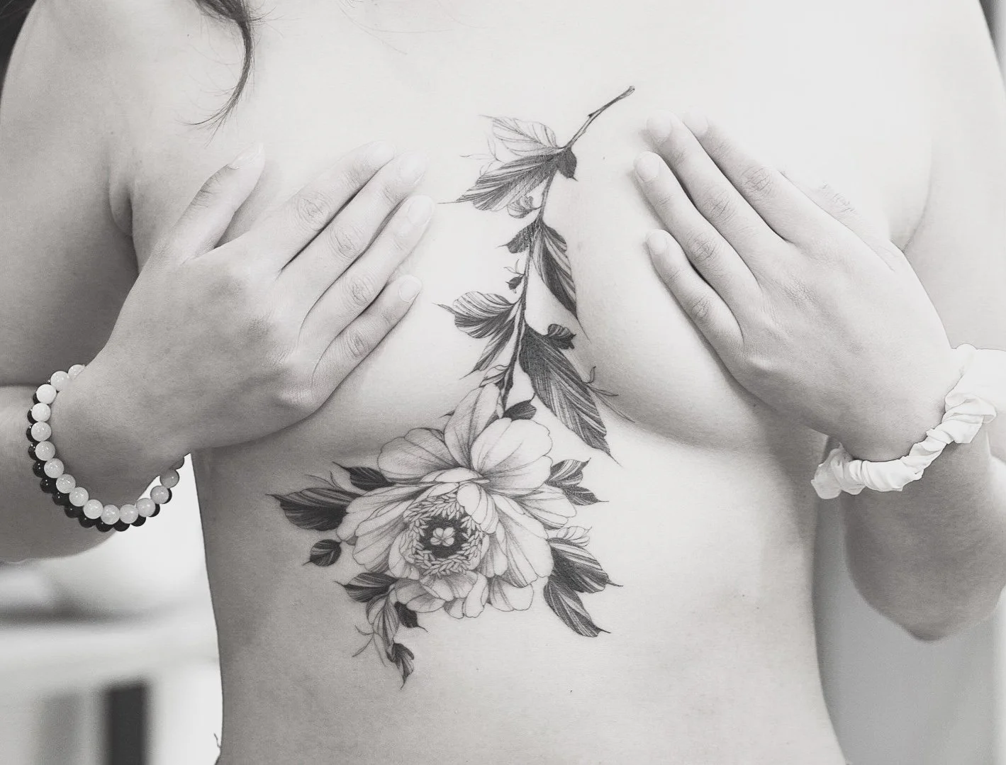 A person with floral tattoos on their chest, covering their breasts with hands, wearing a pearl bracelet on the left wrist and a scrunchie on the right wrist.