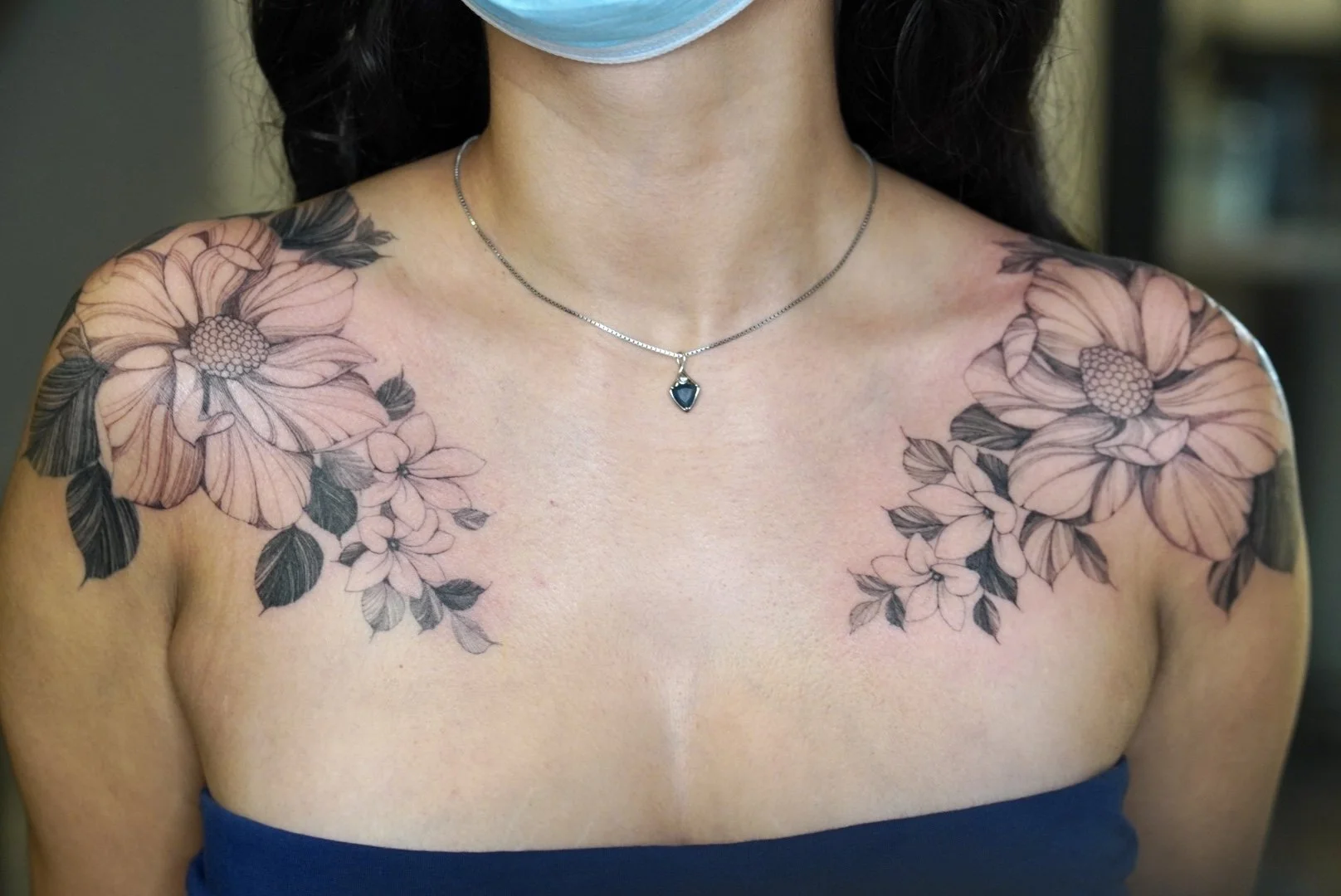 Woman wearing a face mask, a necklace with a blue pendant, and a strapless top, showcasing floral shoulder tattoos.