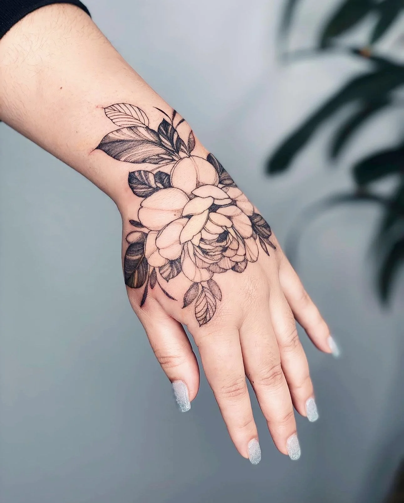 Black and gray floral tattoo on a person's wrist and hand, featuring a large blooming flower with leaves.