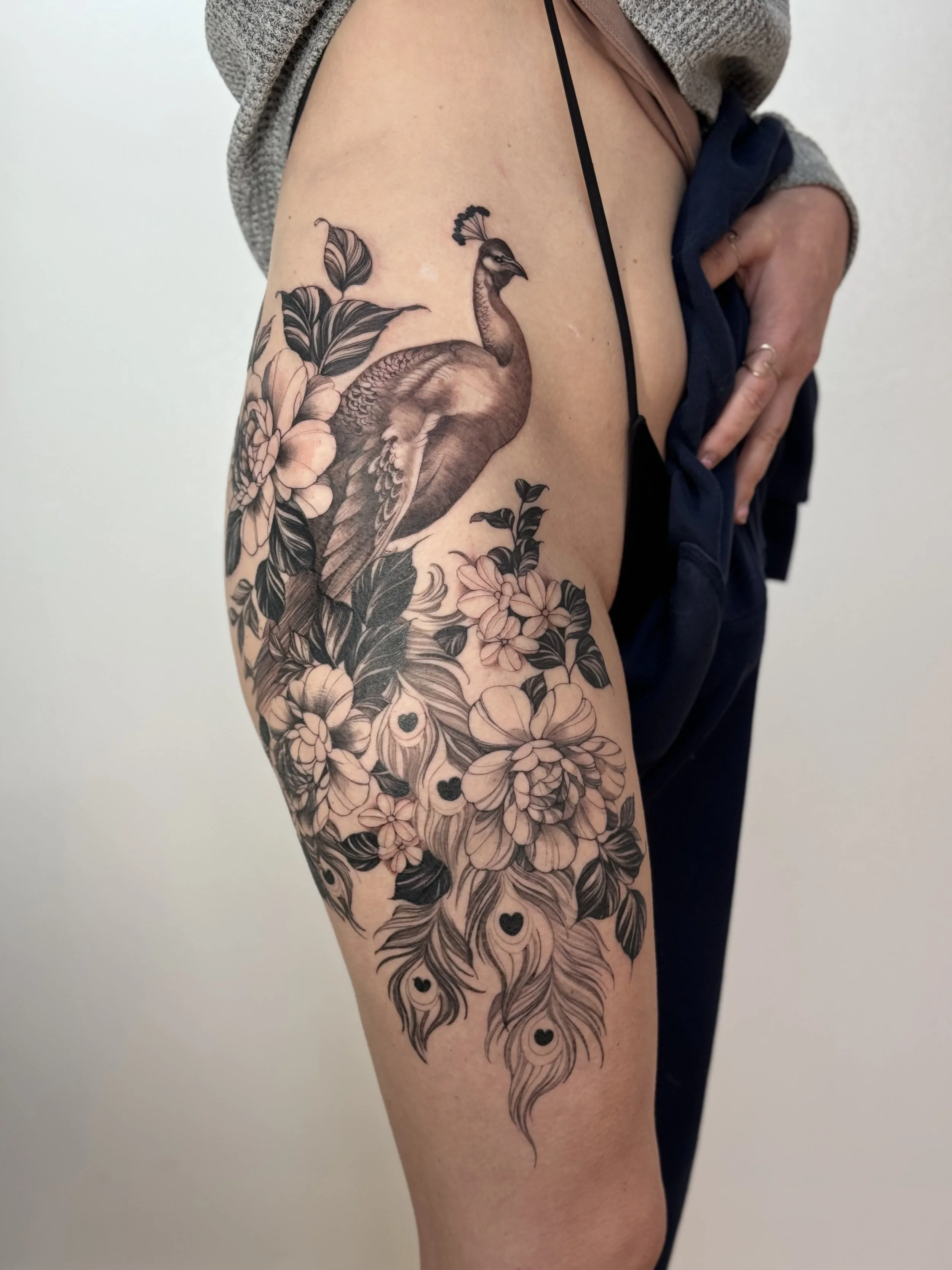 A detailed black and gray tattoo of a peacock surrounded by flowers and leaves on a person's thigh.