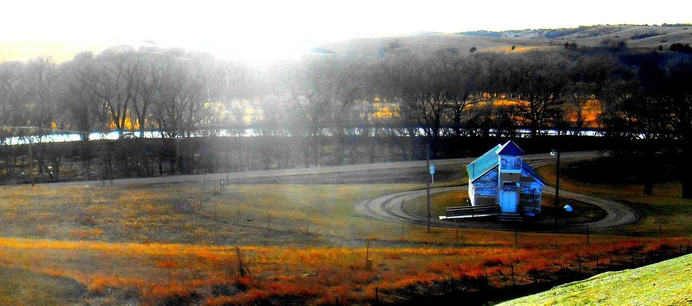 A small blue barn with a curved railway track around it, set in a grassy field with trees in the background during sunset.