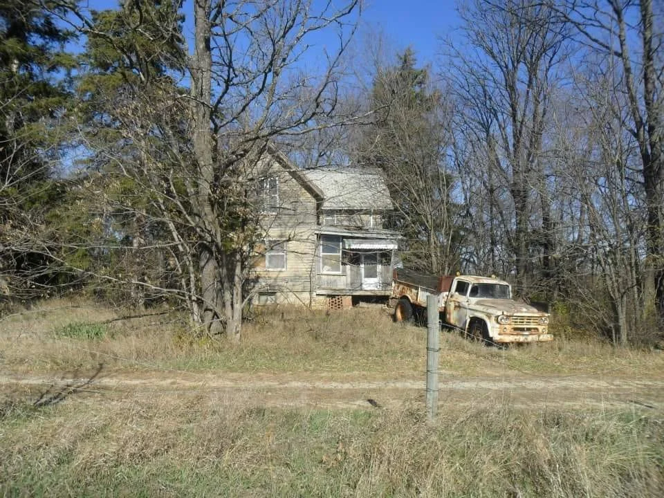 An abandoned house with peeling paint and broken windows, surrounded by leafless trees, and an old rusted pickup truck parked on the grass in front.