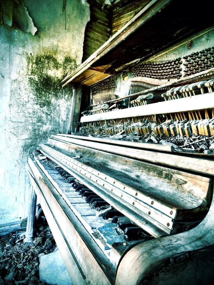 An old, damaged upright piano with broken keys and dust inside a decayed room with peeling walls.