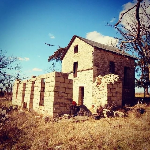 An old stone building with a partially restored front wall, situated in a dry grassy field. The building has two stories with small windows, and there's a leafless tree nearby. A bird is flying in the sky.