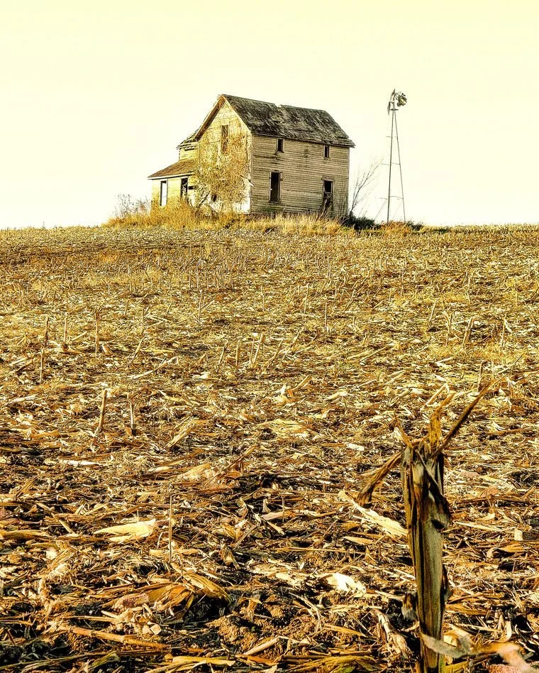 Old abandoned house on a hill with a windmill nearby, surrounded by a harvested cornfield.