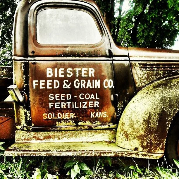 Old rusted truck with faded paint and signage for Biester Feed & Grain Company, advertising seed, coal, and fertilizer, located in Soldier, Kansas, surrounded by green grass and trees.