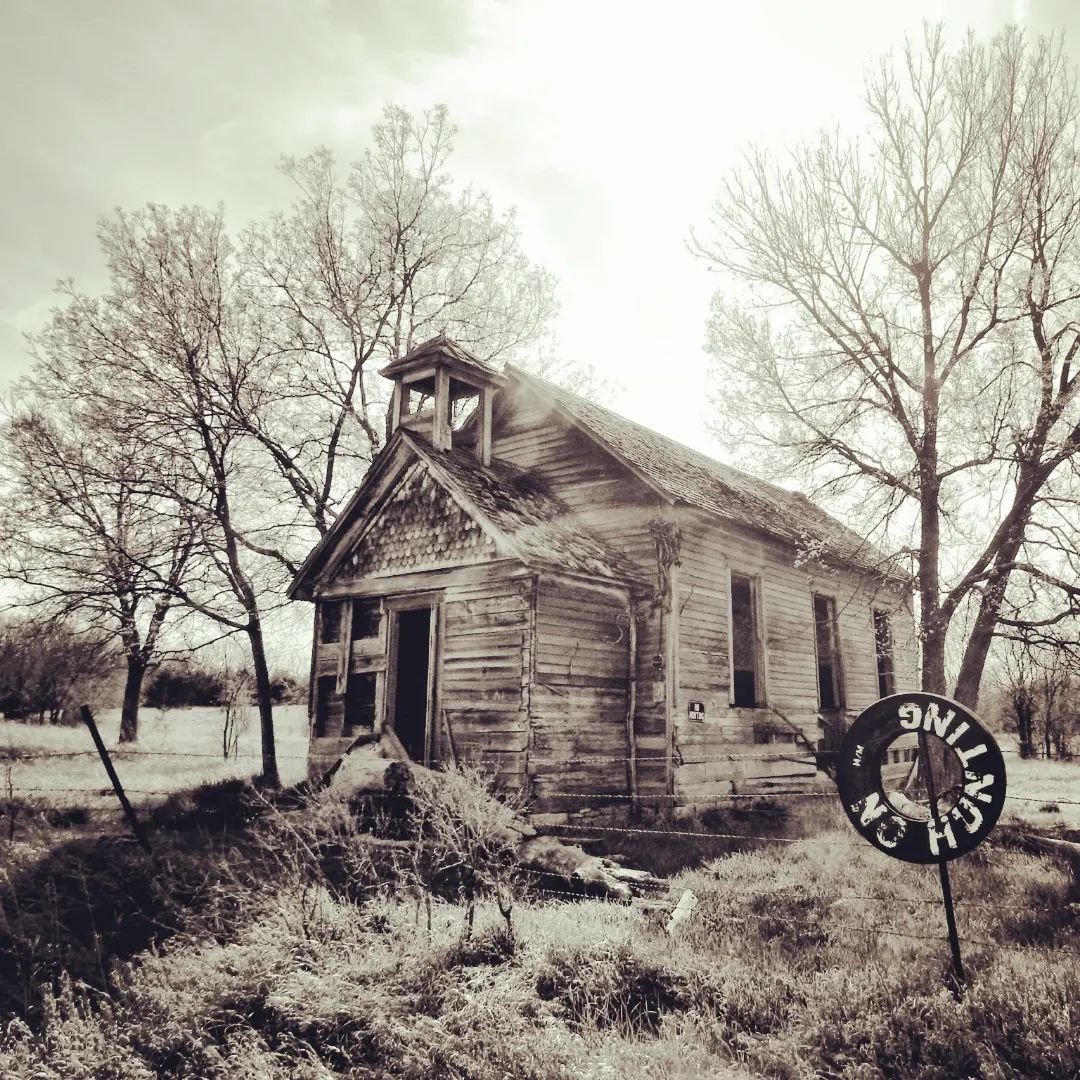 An old, abandoned wooden house with peeling paint, surrounded by leafless trees and overgrown grass, with a damaged fence and a fallen sign that says 'NO TRESPASSING'.