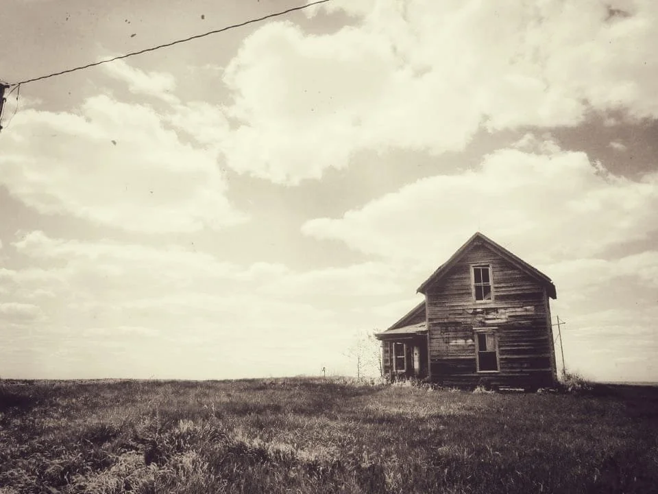 An old, weathered wooden house standing alone in an open field under a cloudy sky.