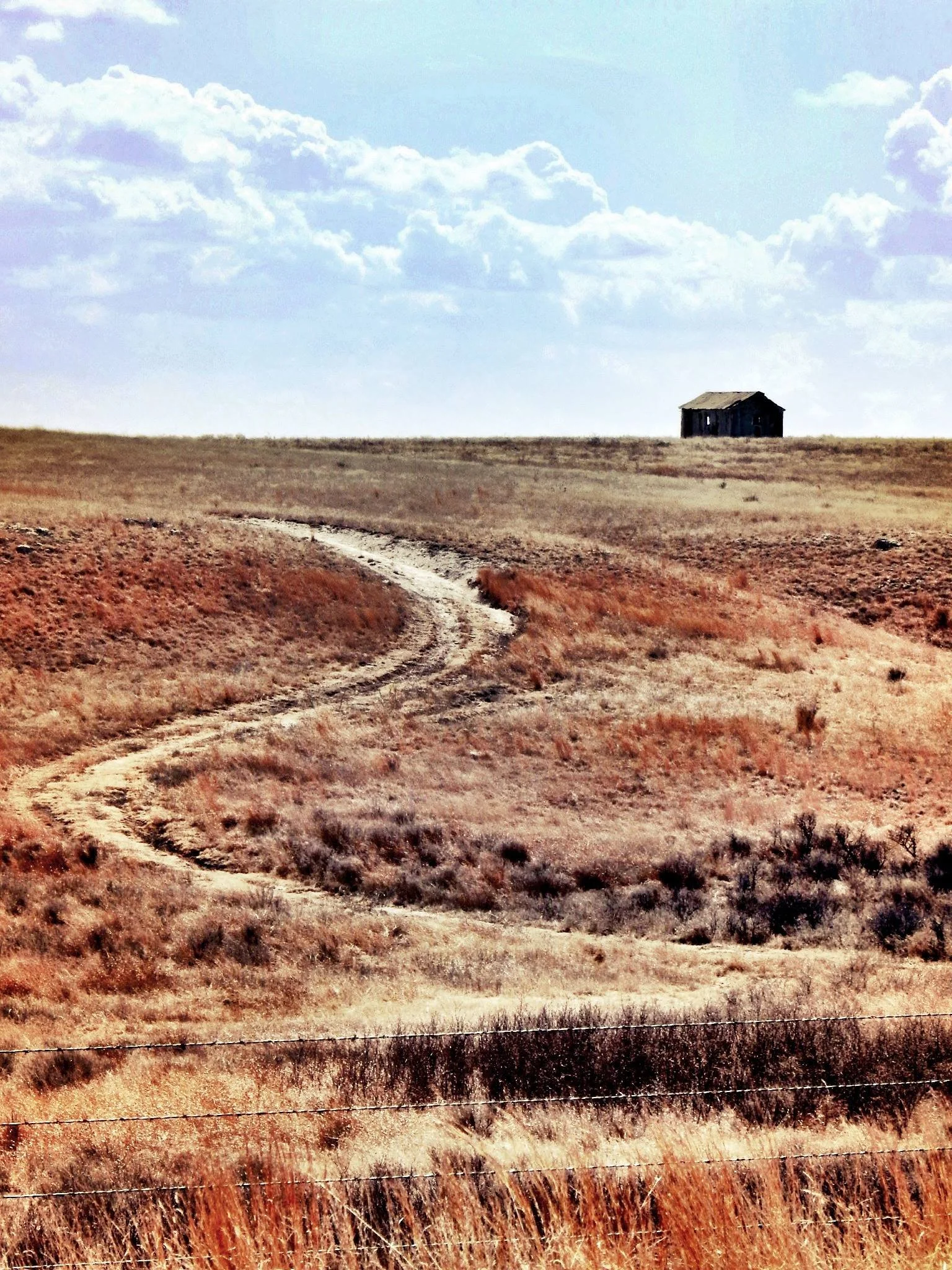 A barren, brown field with a winding dirt path, a barbed wire fence in the foreground, and a small, weathered wooden house on a hill in the background under a partly cloudy sky.